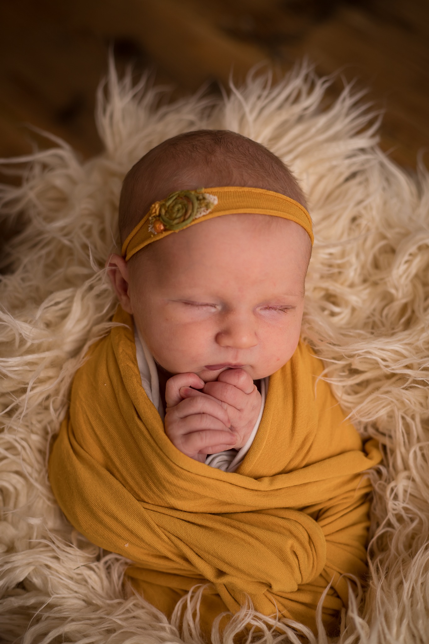 Śpiący noworodek zawinięty w musztardowy materiał, z ozdobną opaską na głowie, leżący na puszystym, kremowym futrze.