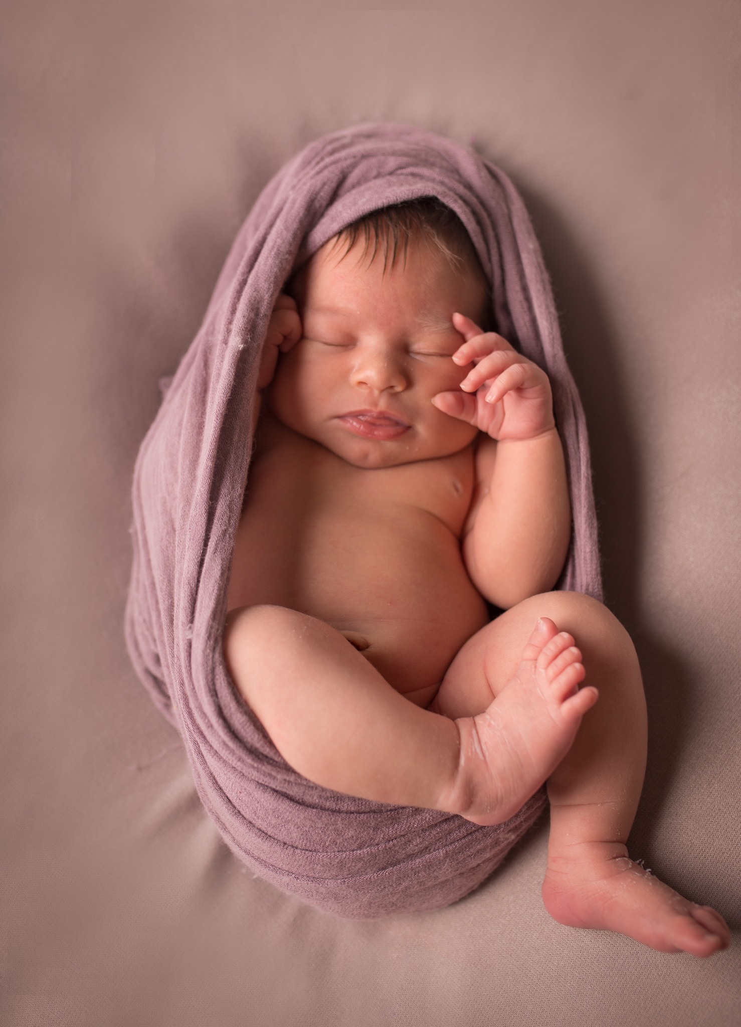 Śpiący noworodek zawinięty w fioletowy materiał na tle w kolorze mauve, z delikatnie uniesioną rączką przy twarzy i widocznymi stópkami.