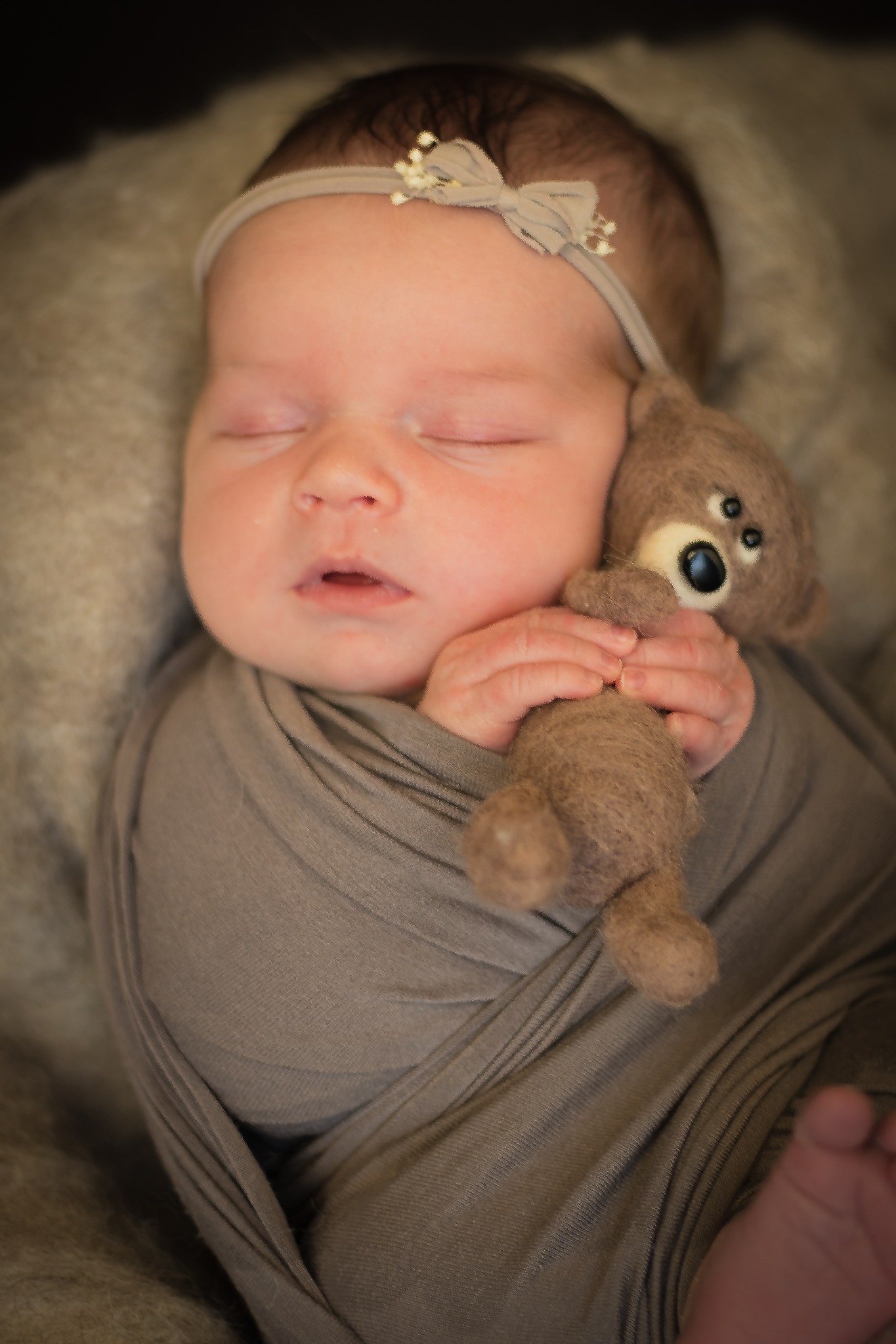 Śpiący noworodek owinięty w szary materiał, z beżową opaską na głowie i pluszowym misiem w rączkach.