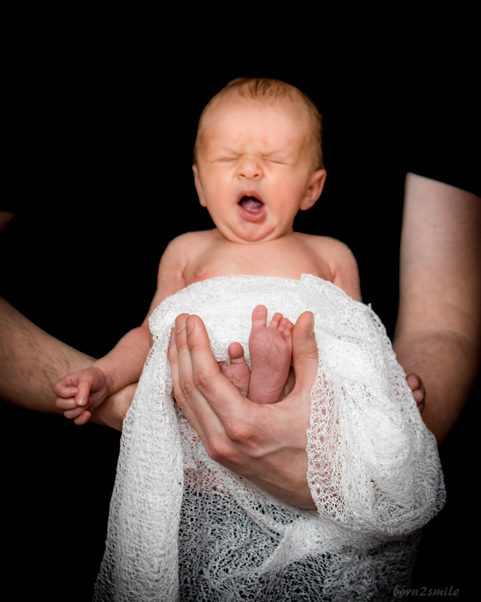 Noworodek ziewający zawinięty w białą, ażurową tkaninę, trzymany w dłoniach na czarnym tle.