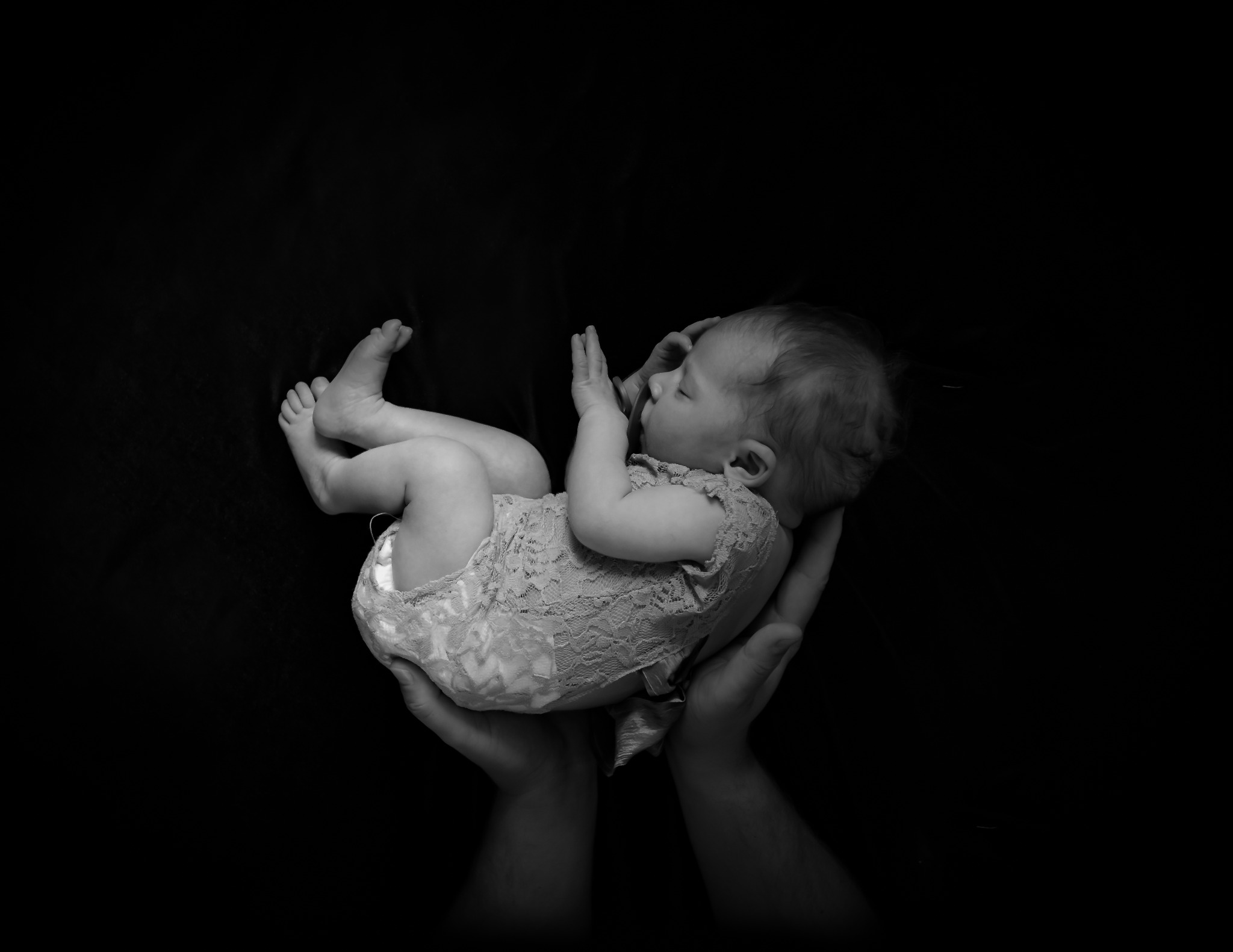 Czarno-białe ujęcie noworodka śpiącego w dłoniach, ubranego w koronkowy strój, na tle ciemnego tła.