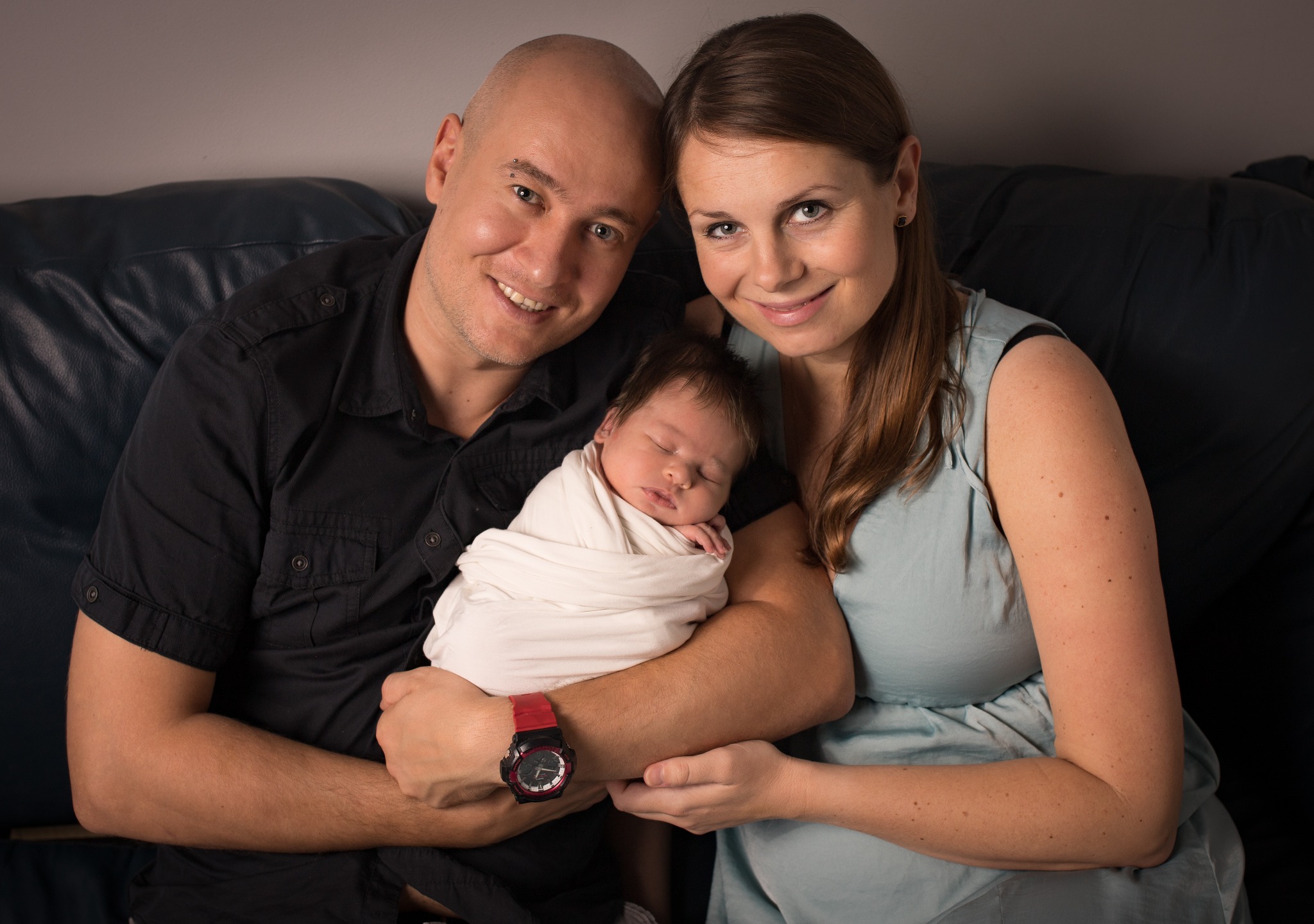 Uśmiechnięta para trzymająca śpiącego noworodka owiniętego w biały kocyk na tle ciemnej skórzanej kanapy. Mężczyzna ma na nadgarstku czerwony zegarek.