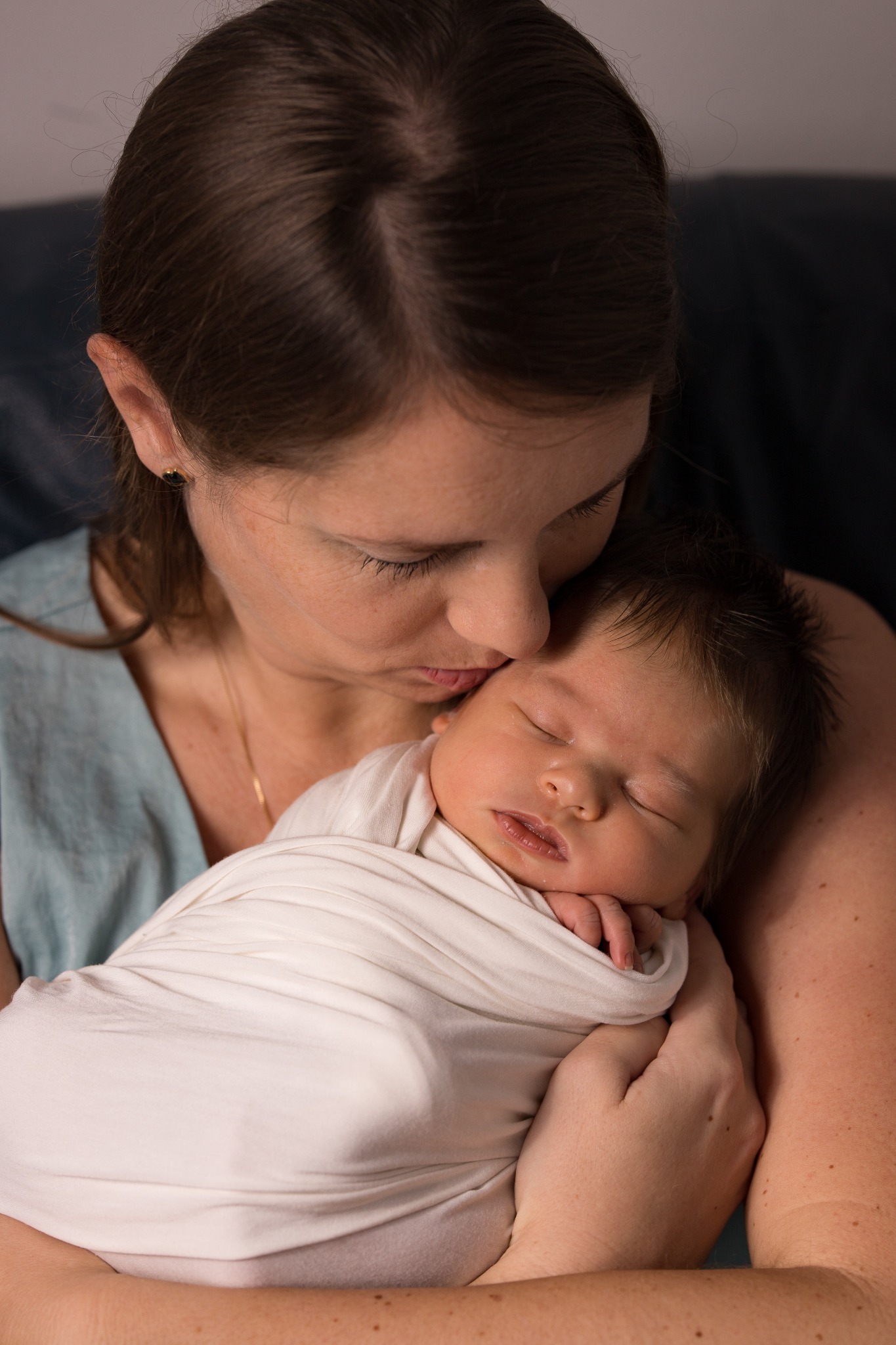 Mama całuje w czoło śpiącego noworodka owiniętego w biały kocyk, widoczne delikatne włoski dziecka i złoty łańcuszek na szyi matki.