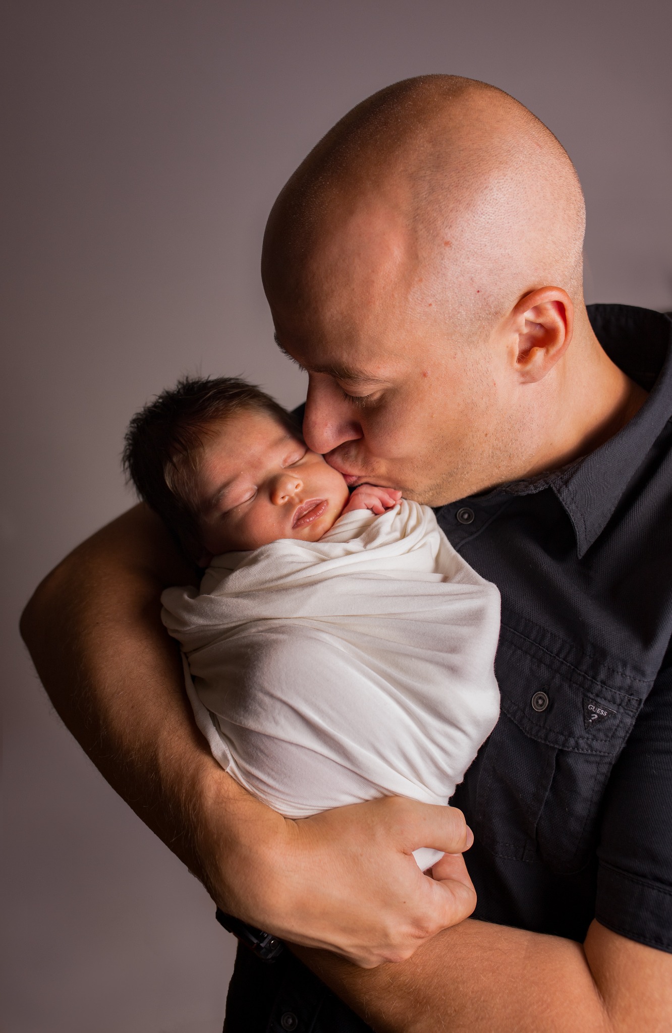 Ojciec czule całuje w policzek noworodka owiniętego w biały kocyk, na jednolitym szarym tle.