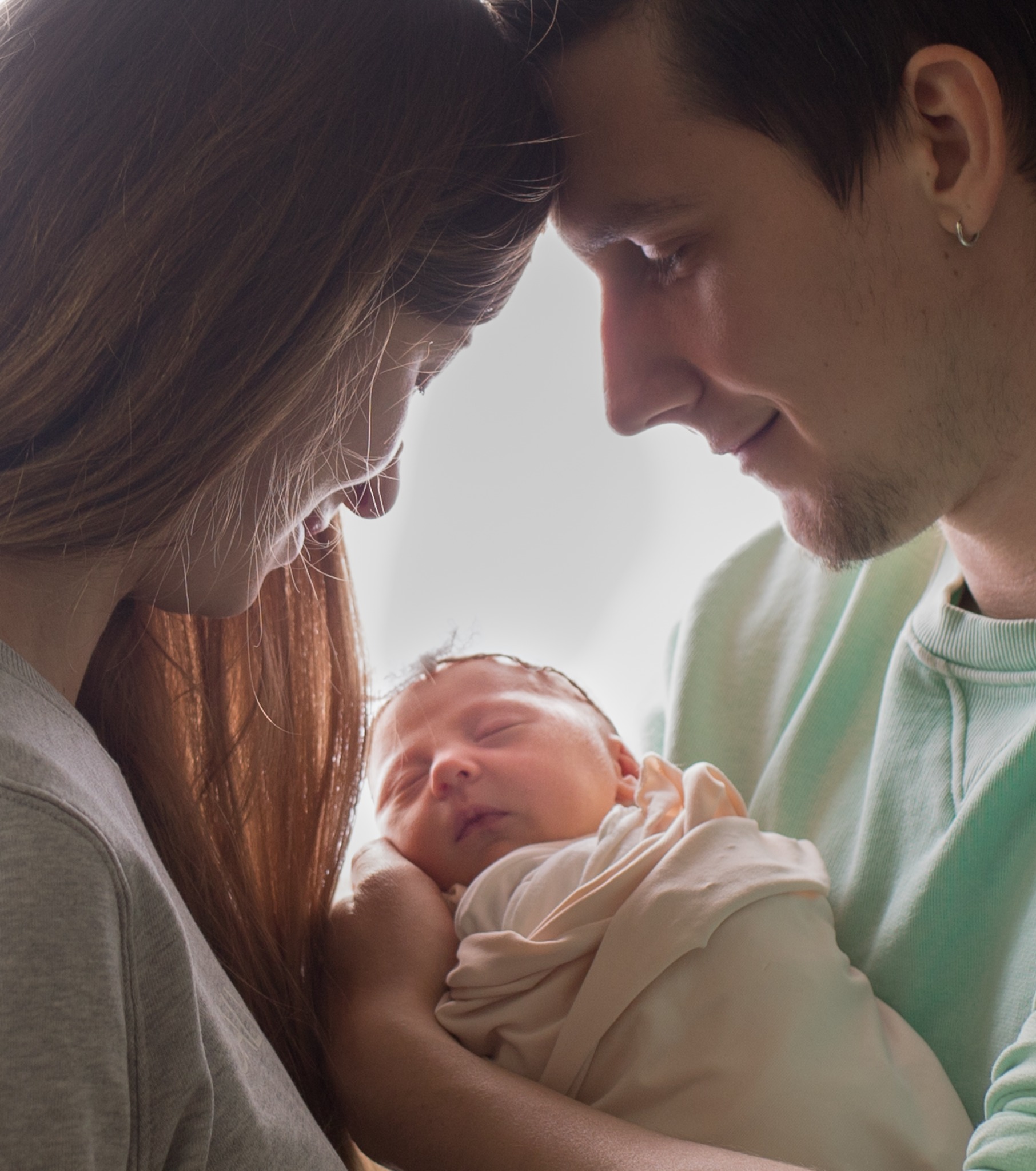 Rodzice spoglądają czule na śpiącego noworodka, owiniętego w jasny materiał, delikatne światło podkreśla bliskość i intymność chwili.