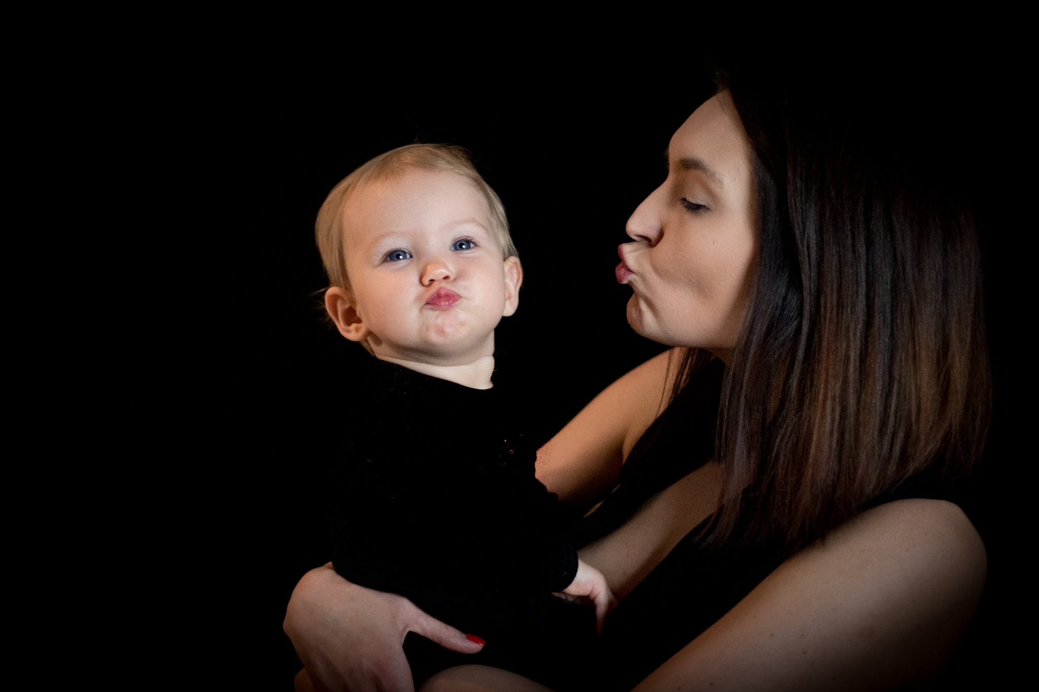 Portret matki z blond dzieckiem na rękach, oboje z ustami ułożonymi w dzióbek na czarnym tle.
