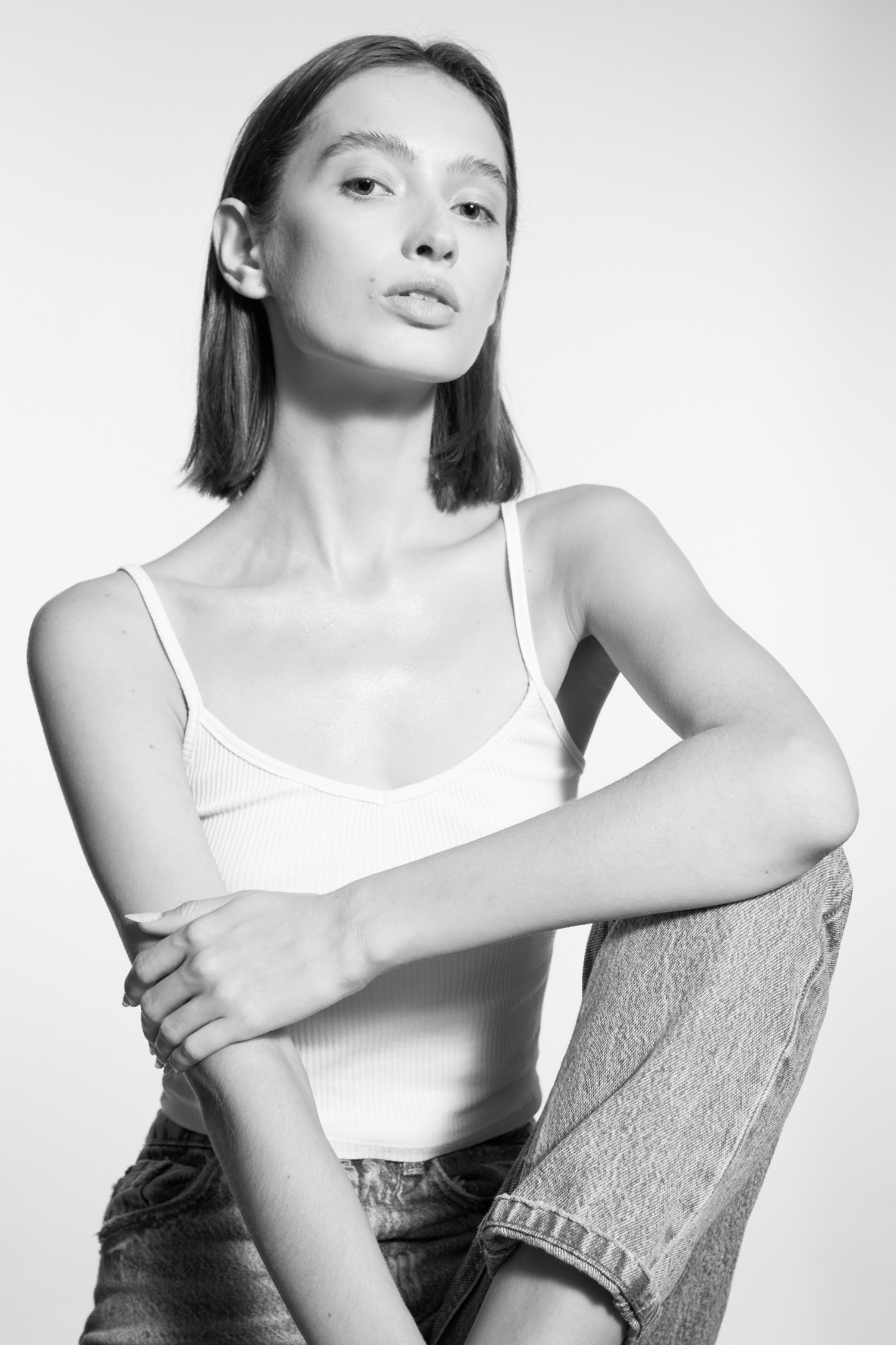 Czarno-białe portretowe ujęcie młodej kobiety w białym topie na ramiączkach i dżinsach, siedzącej z założonymi rękami na kolanach, z delikatnym makijażem i krótkimi włosami.