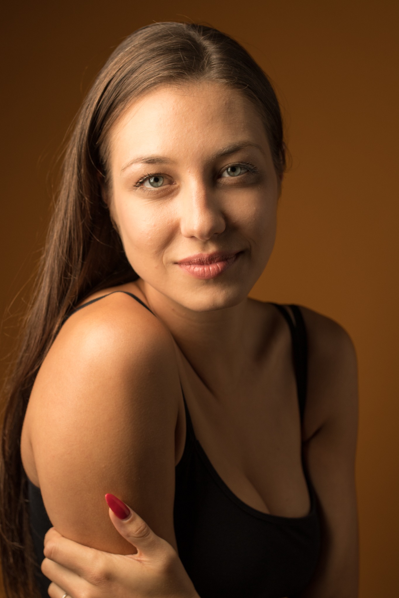 Portret uśmiechniętej kobiety z długimi brązowymi włosami i czerwonymi paznokciami na jednolitym, ciepłym tle.