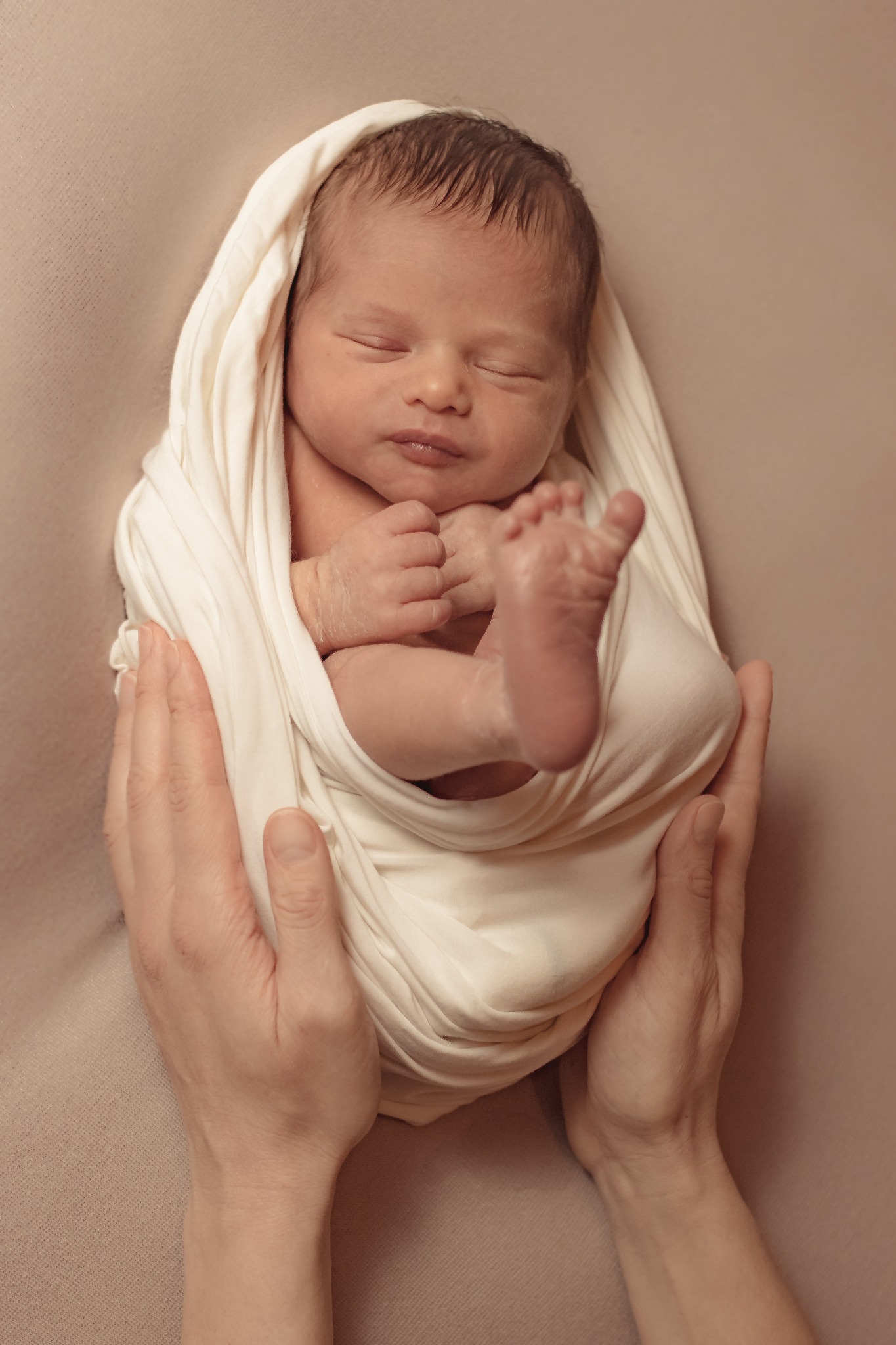 Noworodek owinięty w białą tkaninę, trzymany w dłoniach na beżowym tle, z widoczną małą stópką.