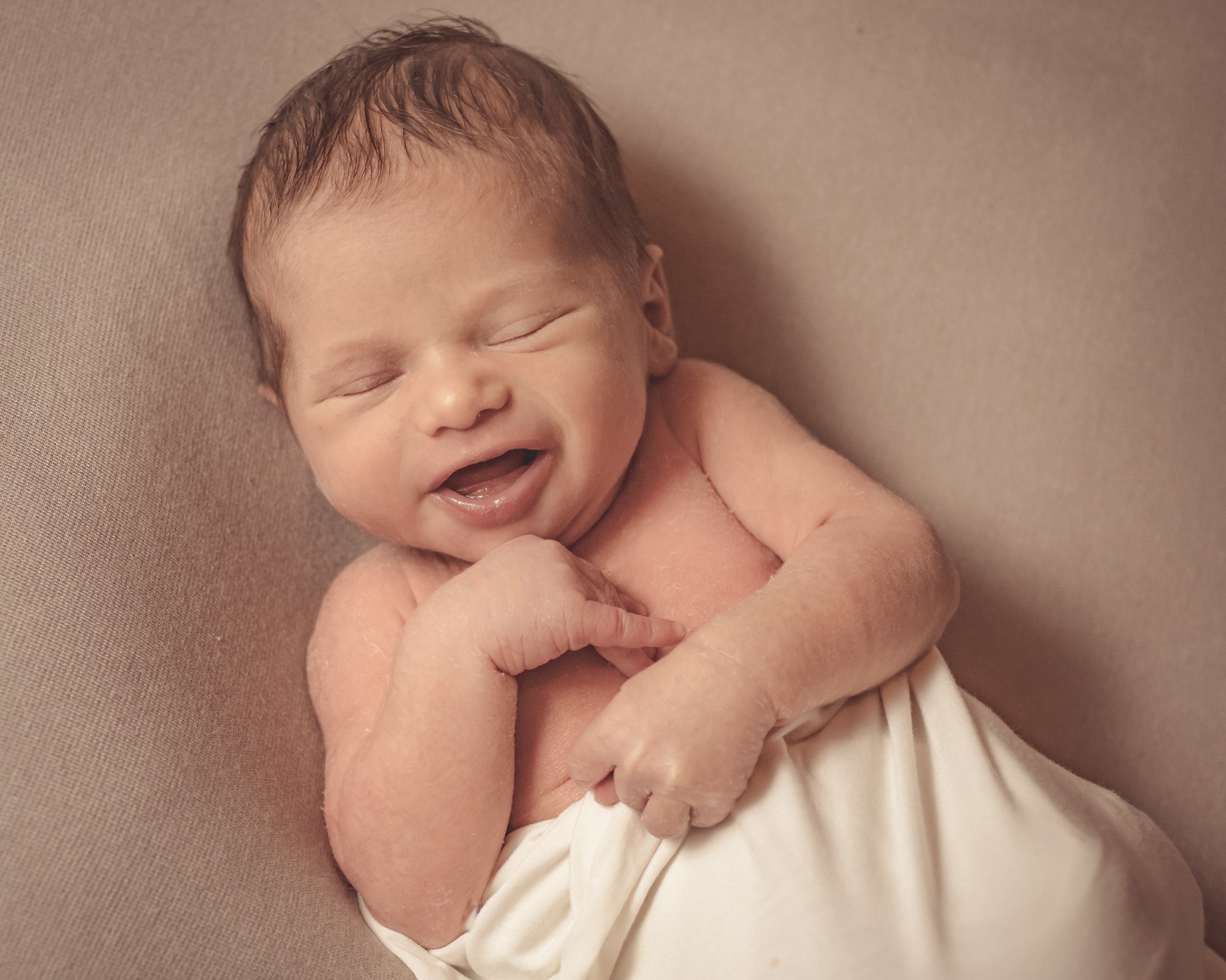 Noworodek leżący na beżowym tle, owinięty w białą tkaninę, z zamkniętymi oczami i lekko otwartymi ustami, uśmiechający się przez sen.