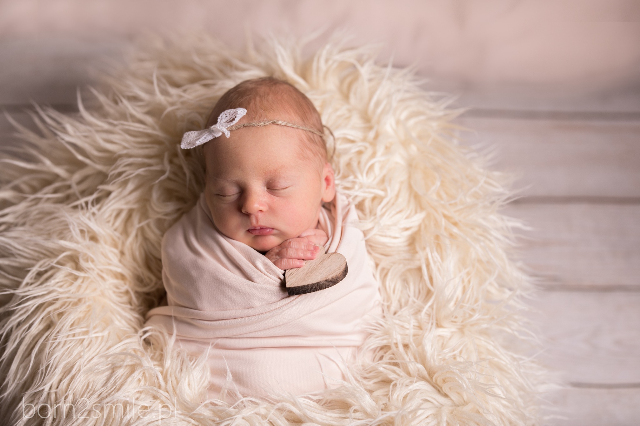 Śpiący noworodek zawinięty w jasnoróżowy materiał, z drewnianym sercem i opaską z kokardką, leży na puszystym, kremowym futrze na tle drewnianych desek.