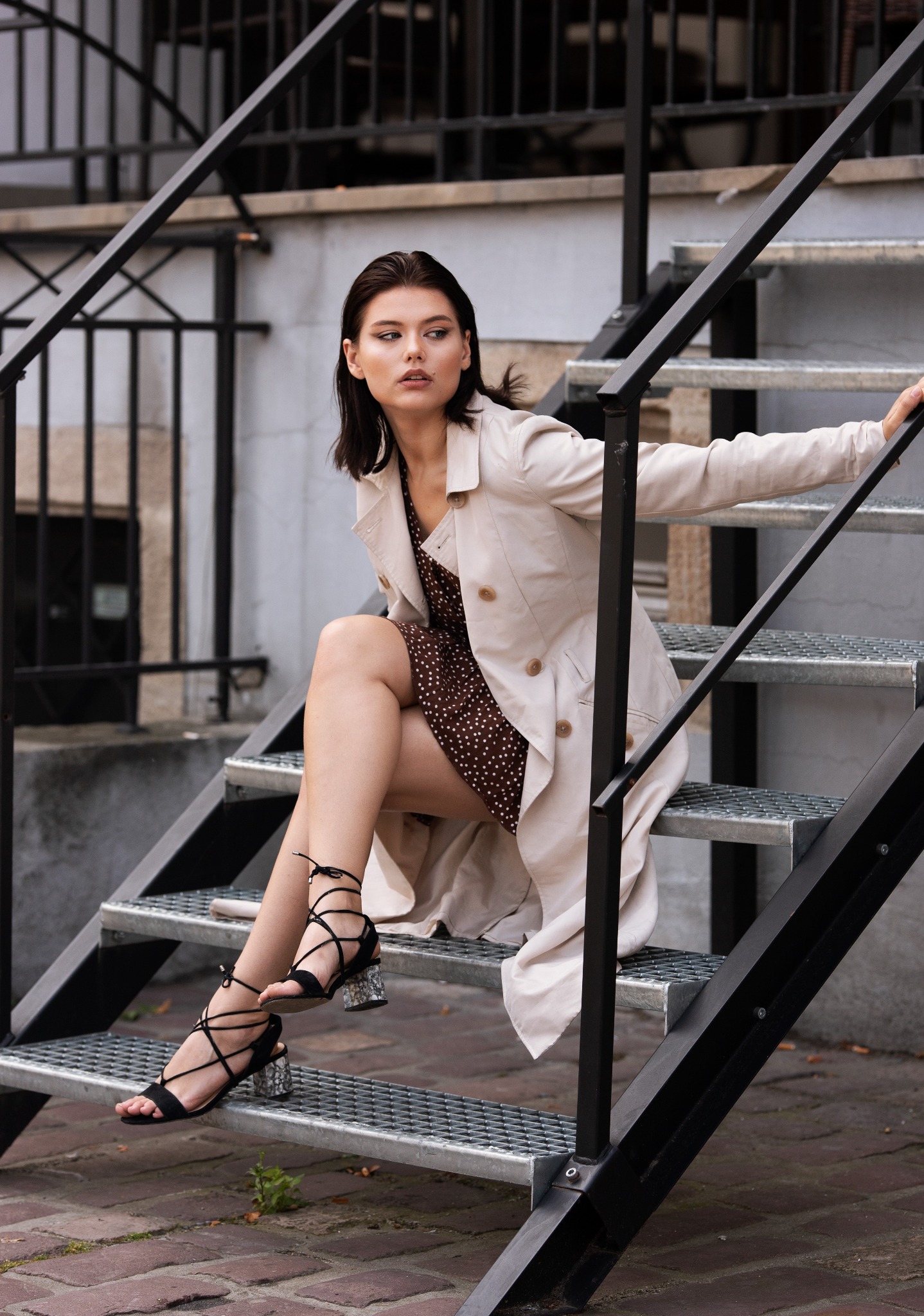 Kobieta w beżowym płaszczu i brązowej sukience w kropki siedzi na metalowych schodach zewnętrznych, pozując w czarnych sandałach na obcasie