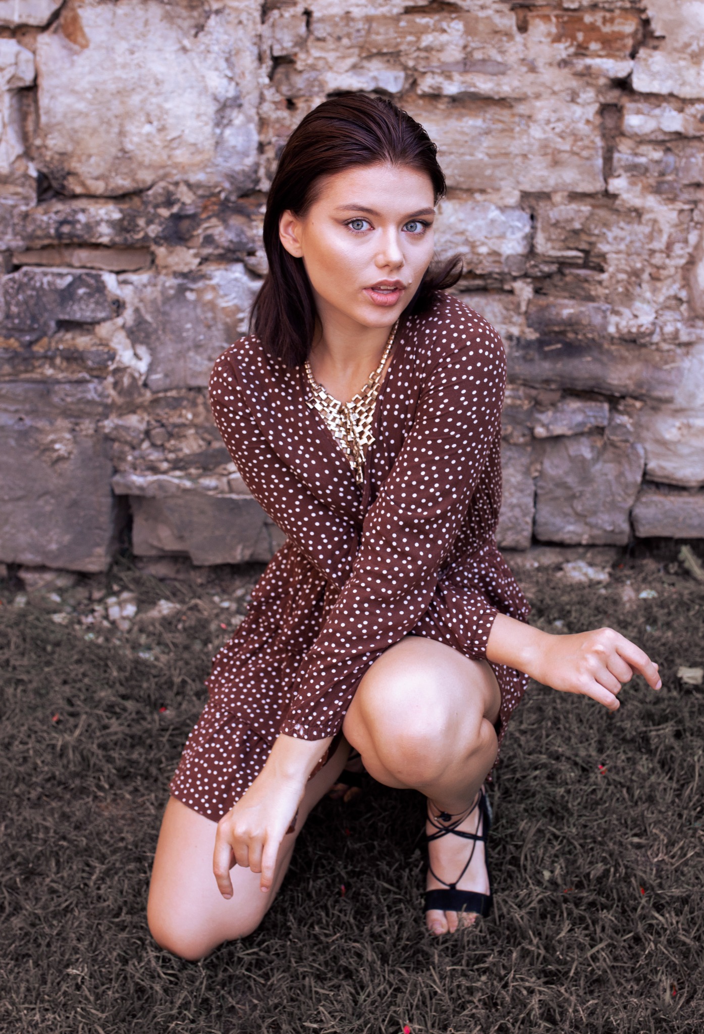 Kobieta w brązowej sukience w białe kropki, klęcząca na trawie przed kamienną ścianą, z grubym złotym łańcuchem na szyi i czarnych sandałach na obcasie.