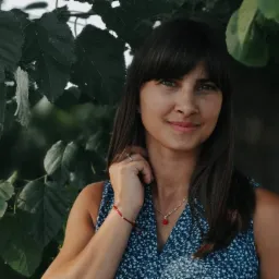 Portret uśmiechniętej brunetki z grzywką, w niebieskiej sukience w kwiaty, z czerwoną bransoletką i naszyjnikiem z koralem na tle zielonych liści.