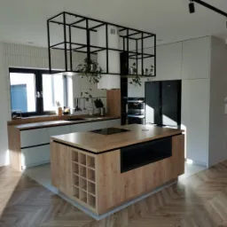 Nowoczesna kuchnia z wyspą, drewnianym blatem i czarną metalową konstrukcją sufitową z roślinami, parkiet w jodełkę, minimalistyczne szafki w odcieniach szarości.