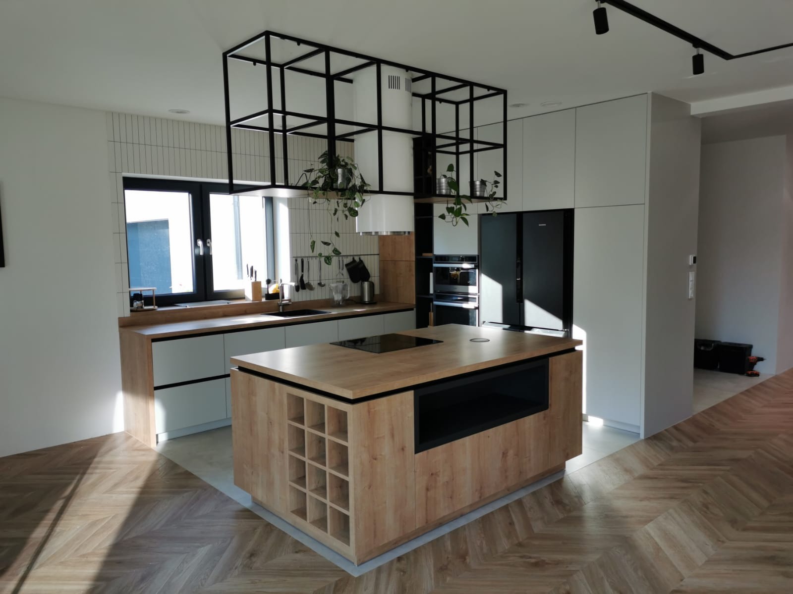 Nowoczesna kuchnia z wyspą, drewnianym blatem i czarną metalową konstrukcją sufitową z roślinami, parkiet w jodełkę, minimalistyczne szafki w odcieniach szarości.
