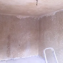 Przygotowanie ścian do malowania w Węgorzewie: ściany pokryte wstępną warstwą gładzi gipsowej z widocznymi śladami szpachlowania, drabina oparta o ścianę w rogu pokoju, surowy stan wykończenia.