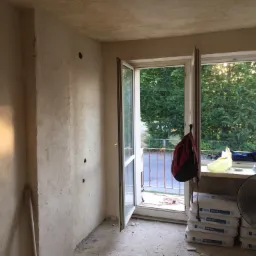 Pokój w trakcie prac wykończeniowych: surowe ściany z widocznymi śladami gładzi, otwarte okno balkonowe z widokiem na zieleń, worki z zaprawą Knauf MP 75 L i wentylator w rogu pomieszczenia.