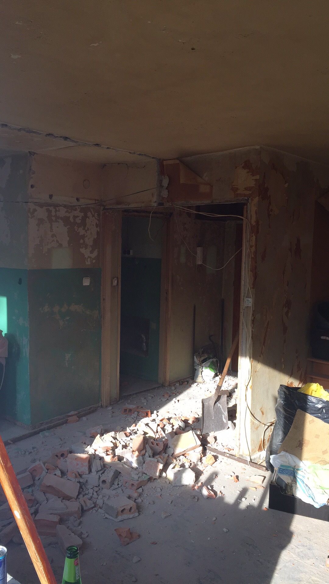 Wnętrze w trakcie demontażu ścian, widoczne gruzowisko z cegieł i odsłonięte przewody elektryczne, ściany z resztkami farby i tapet, światło słoneczne podkreśla bałagan.