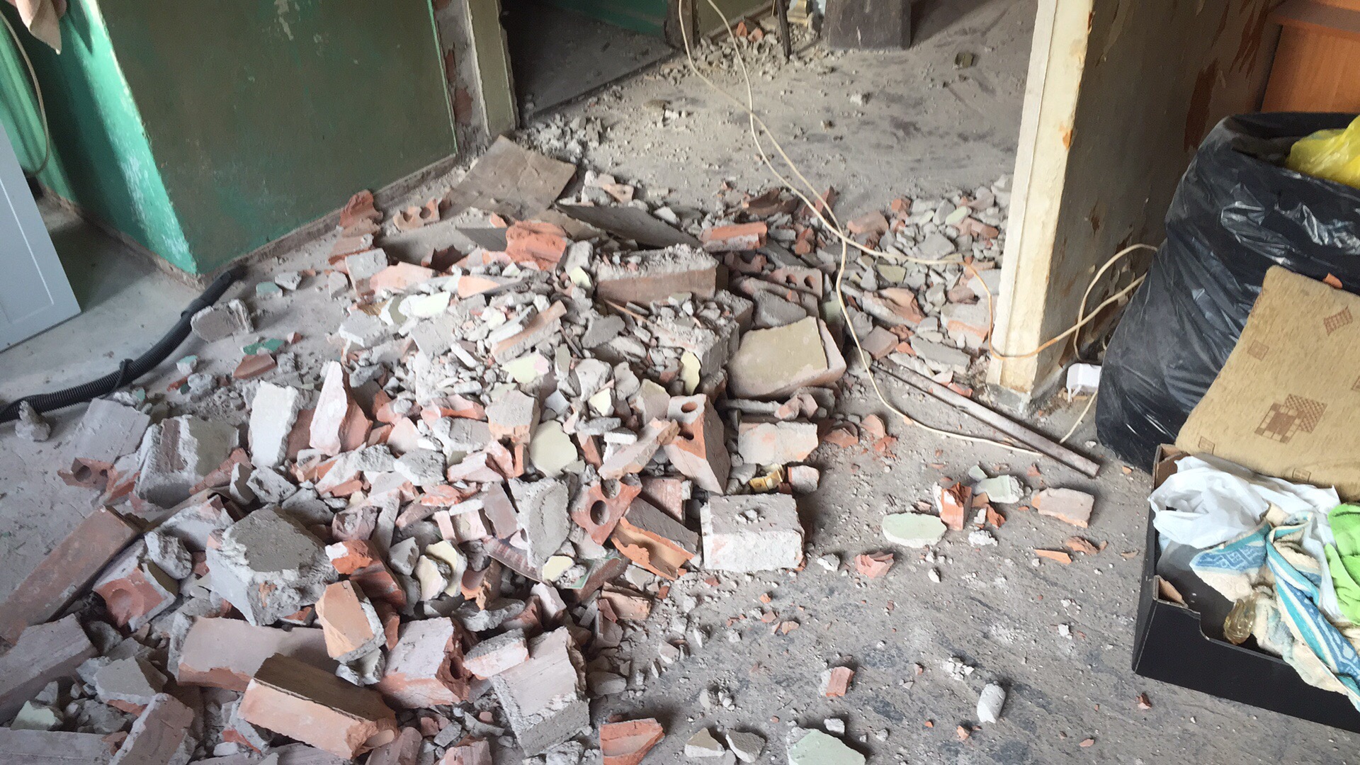 Gruz ceglany i betonowy na podłodze w trakcie demontażu ściany podczas prac remontowych, widoczna zielona ściana w tle i elementy wyposażenia zabezpieczone w workach.