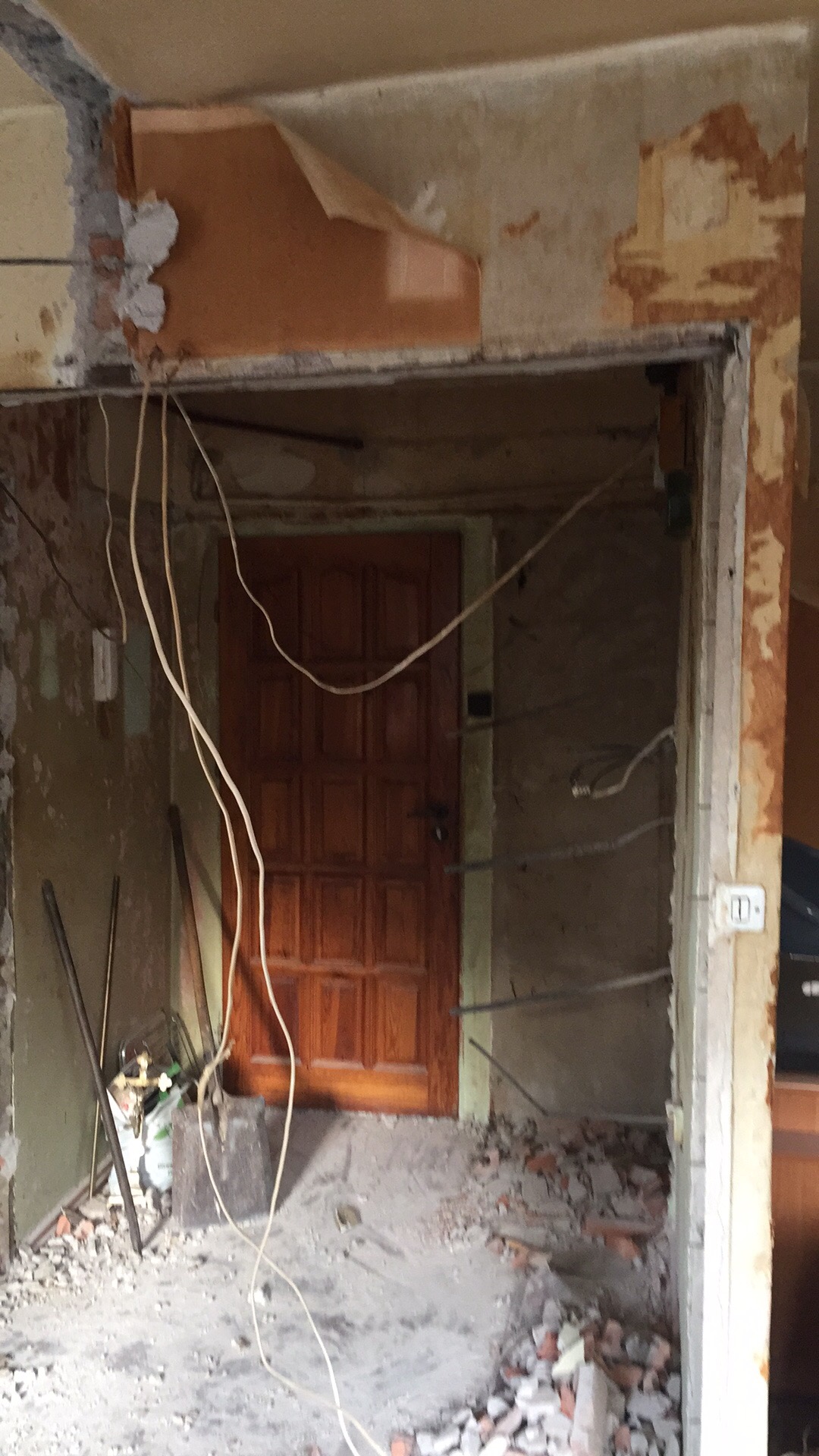 Wnętrze w trakcie demontażu: zerwana tapeta, wystające przewody elektryczne, gruz na podłodze, widoczna drewniana ościeżnica i drzwi.