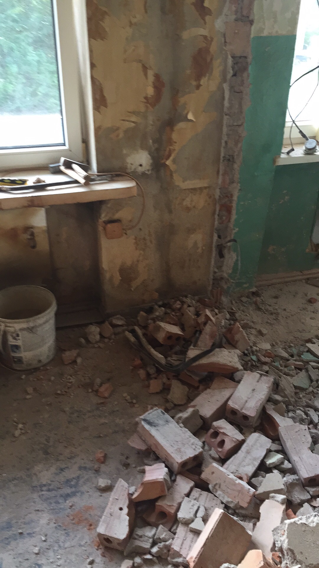 Demontaż ściany w trakcie prac remontowych: widoczne odsłonięte cegły, resztki tapety na ścianach, okno z parapetem i wiadro po farbie.