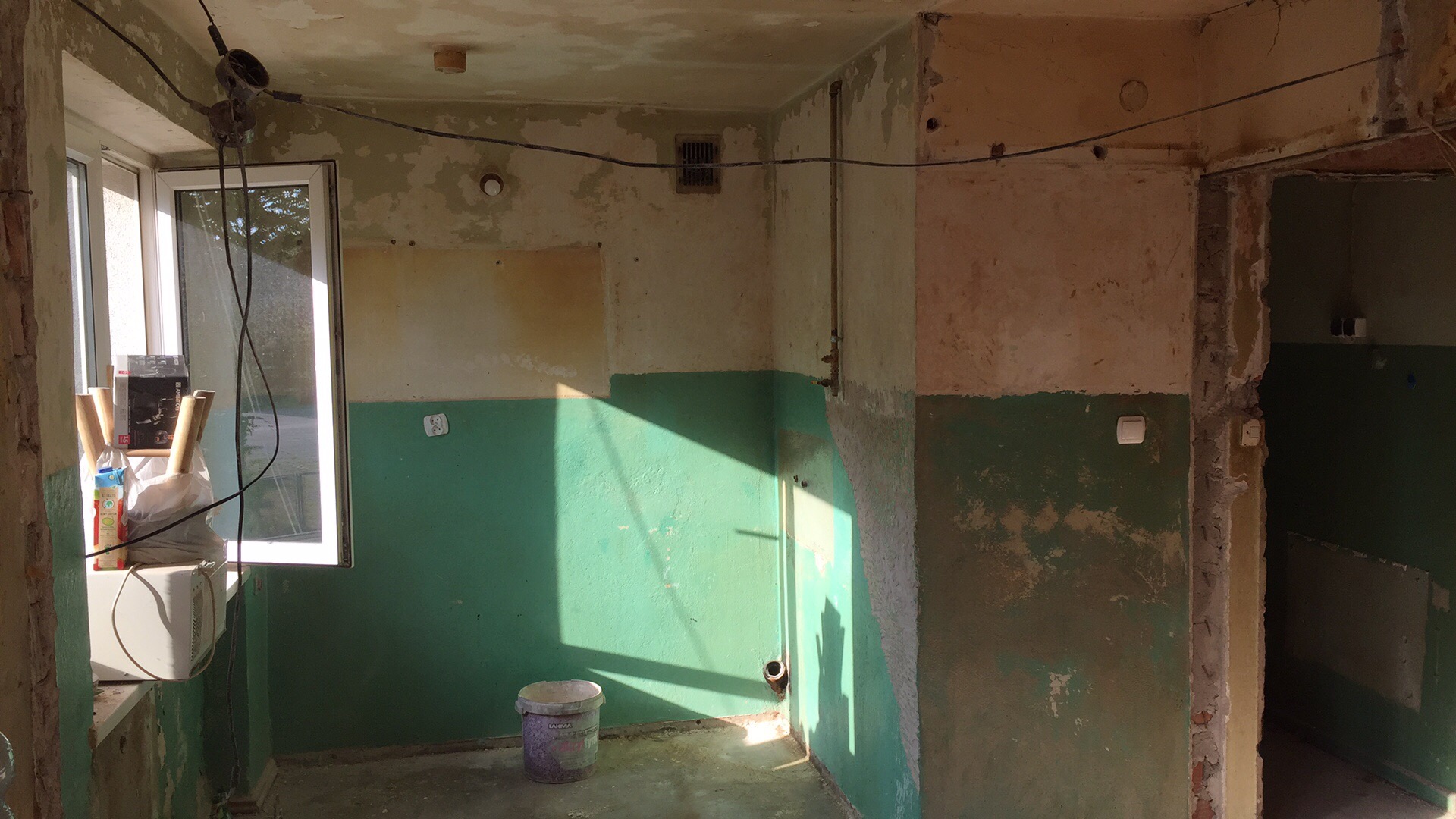 Wnętrze pomieszczenia w trakcie demontażu, widoczne fragmenty starej farby, odsłonięte przewody elektryczne, okno z materiałami budowlanymi, kubeł na podłodze, ślady po instalacjach na ścianach.