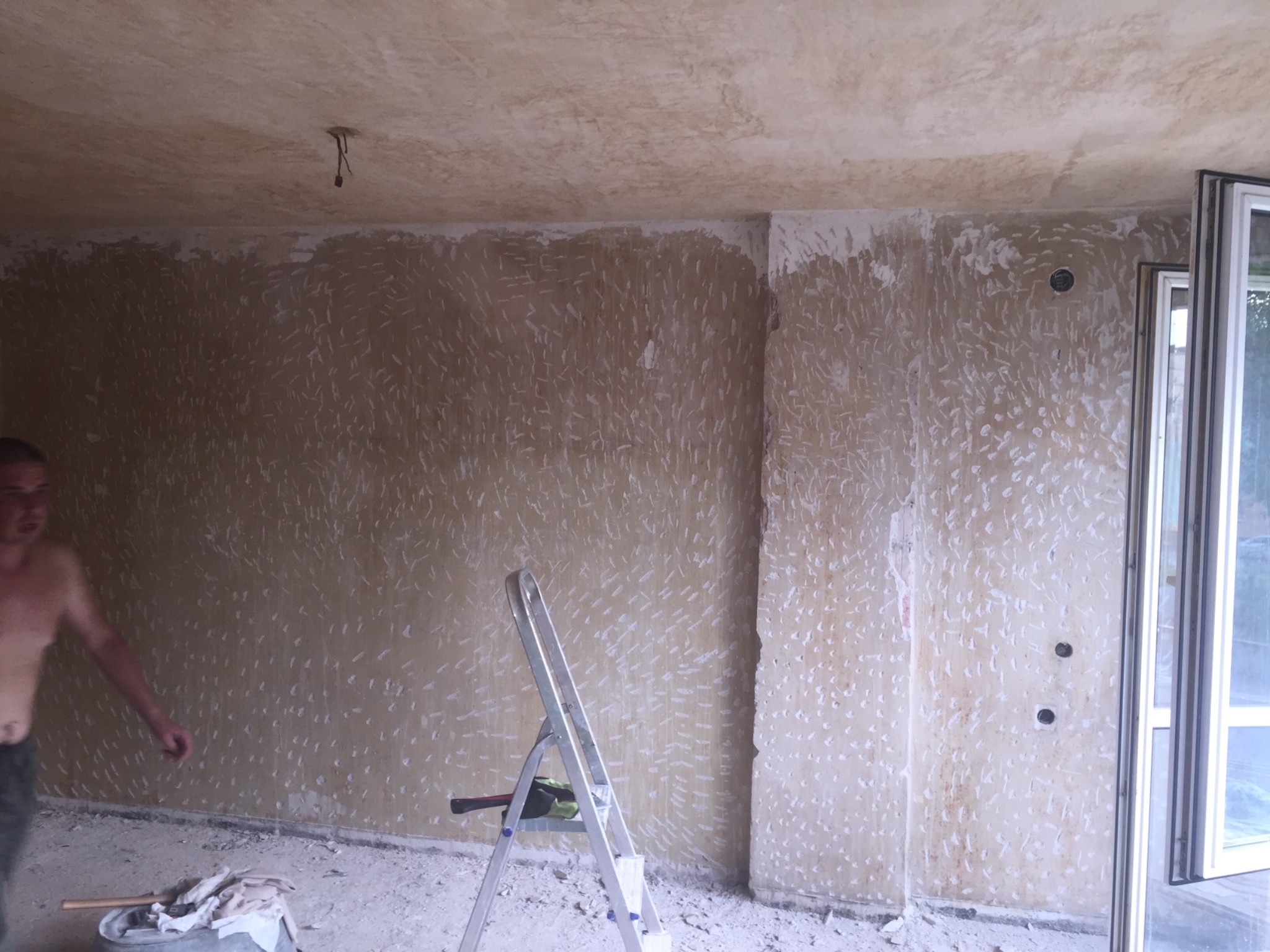 Przygotowanie ścian do malowania: ściany pokryte gładzią szpachlową, widoczna drabina, narzędzia i fragment sylwetki osoby w trakcie pracy.