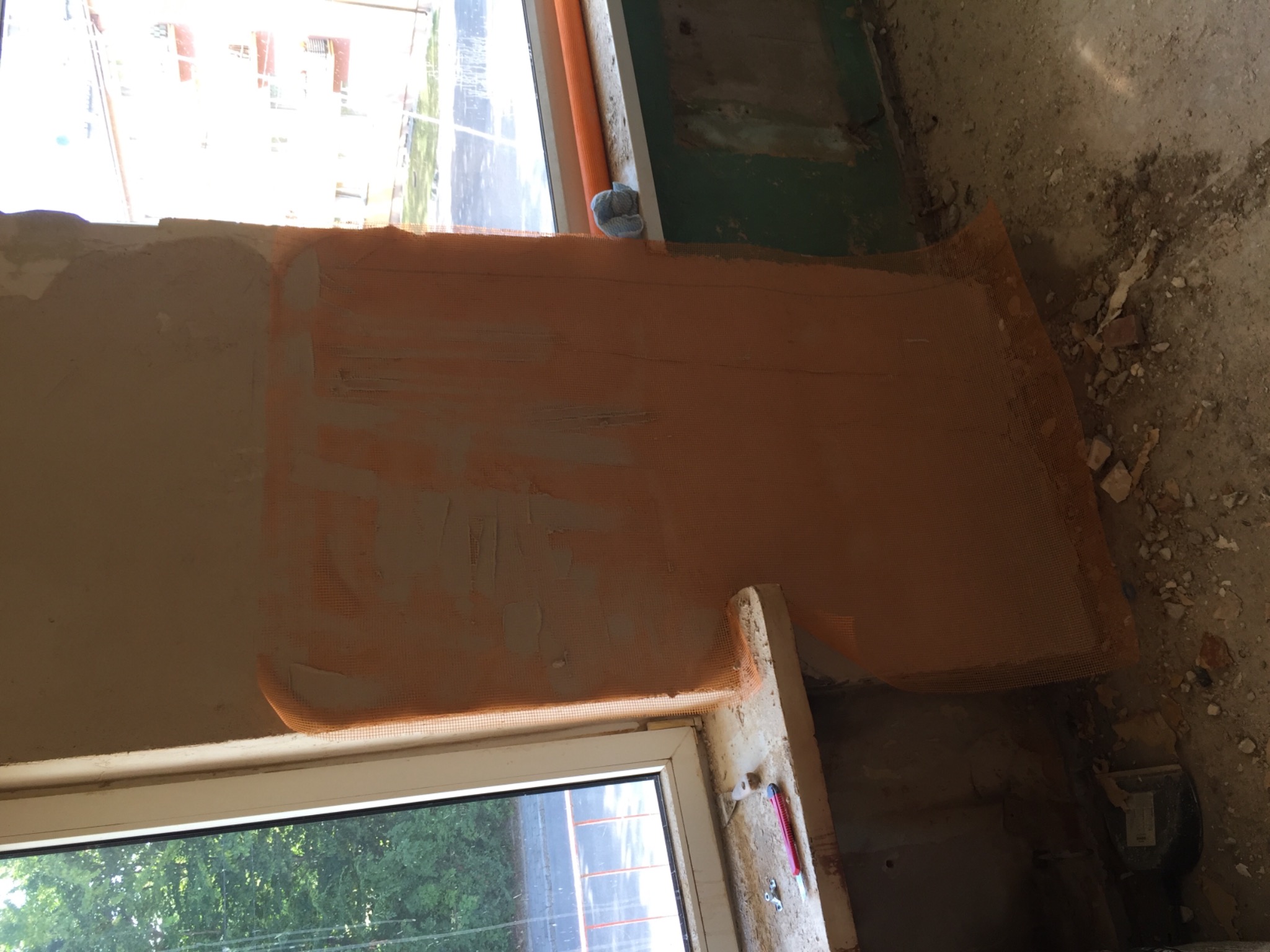 Ściana z nałożoną siatką wzmacniającą, przygotowana do tynkowania, widoczny fragment okna i zabrudzona podłoga w trakcie prac.