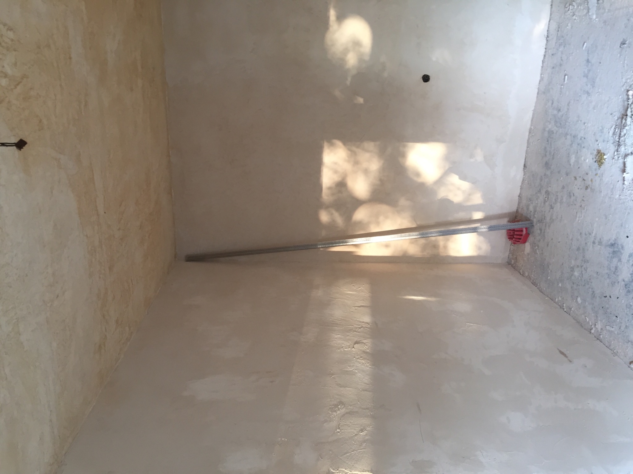 Pokój w trakcie szpachlowania ścian, widoczne ślady gładzi, metalowa listwa montażowa i plama światła słonecznego na ścianie.