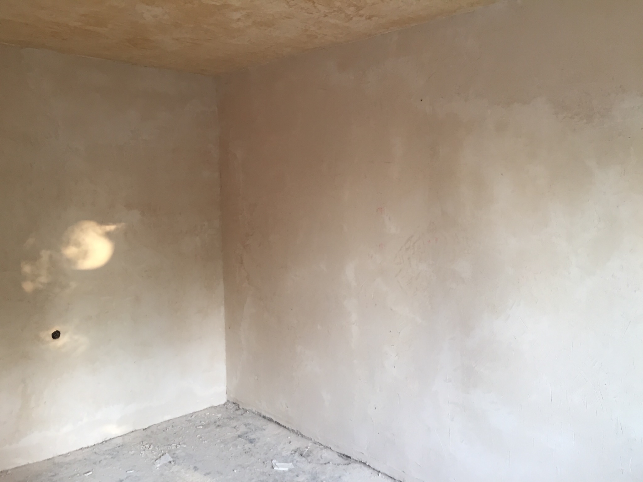 Surowe ściany i sufit w rogu pokoju po nałożeniu gładzi, widoczne nierówności i niedoskonałości przed malowaniem, oraz otwór na gniazdko elektryczne.