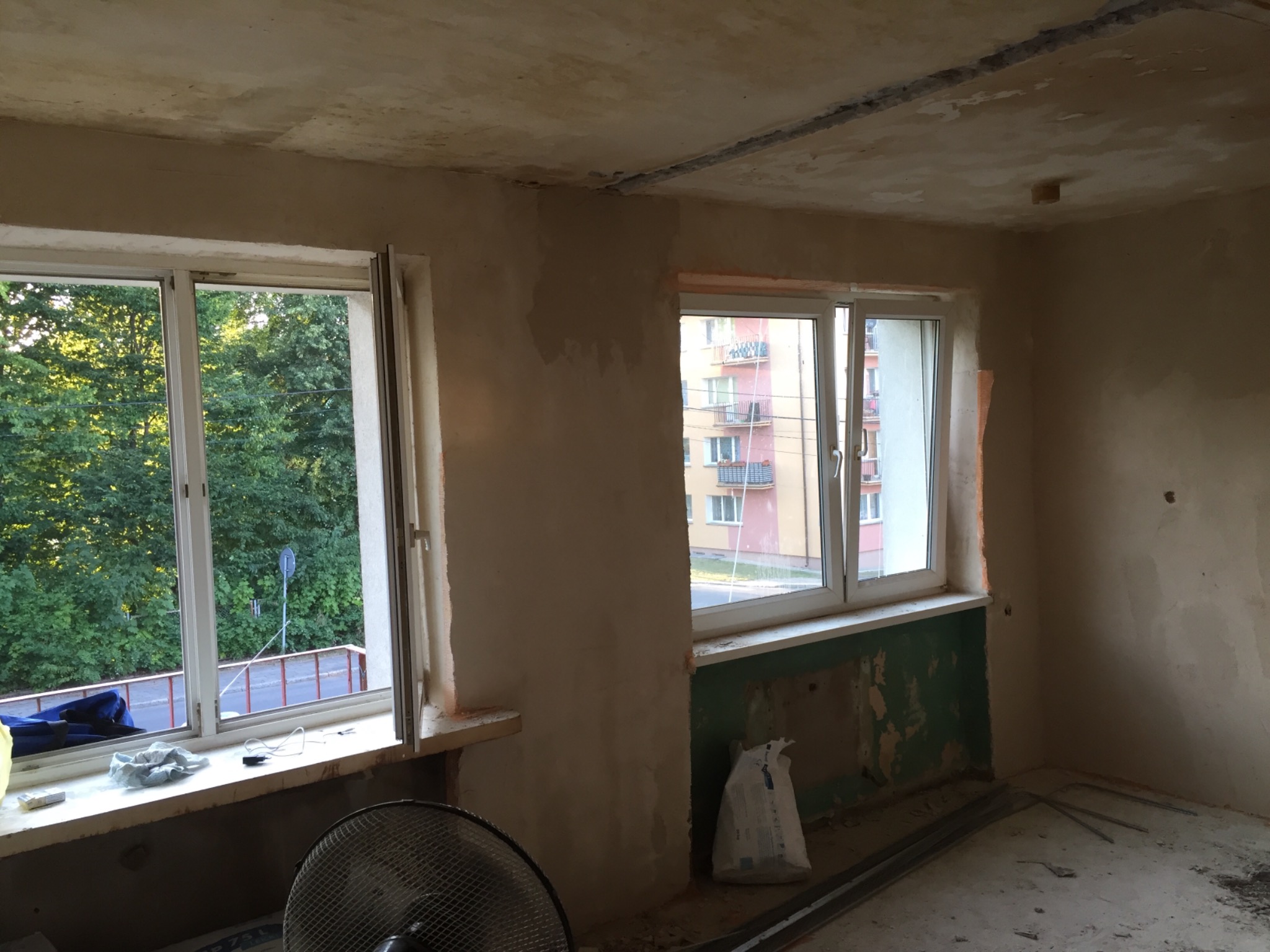 Pokój w trakcie prac przygotowawczych do wykończenia, ściany pokryte gładzią, dwa okna, widoczne narzędzia i materiały budowlane, w tle budynki mieszkalne i zieleń.