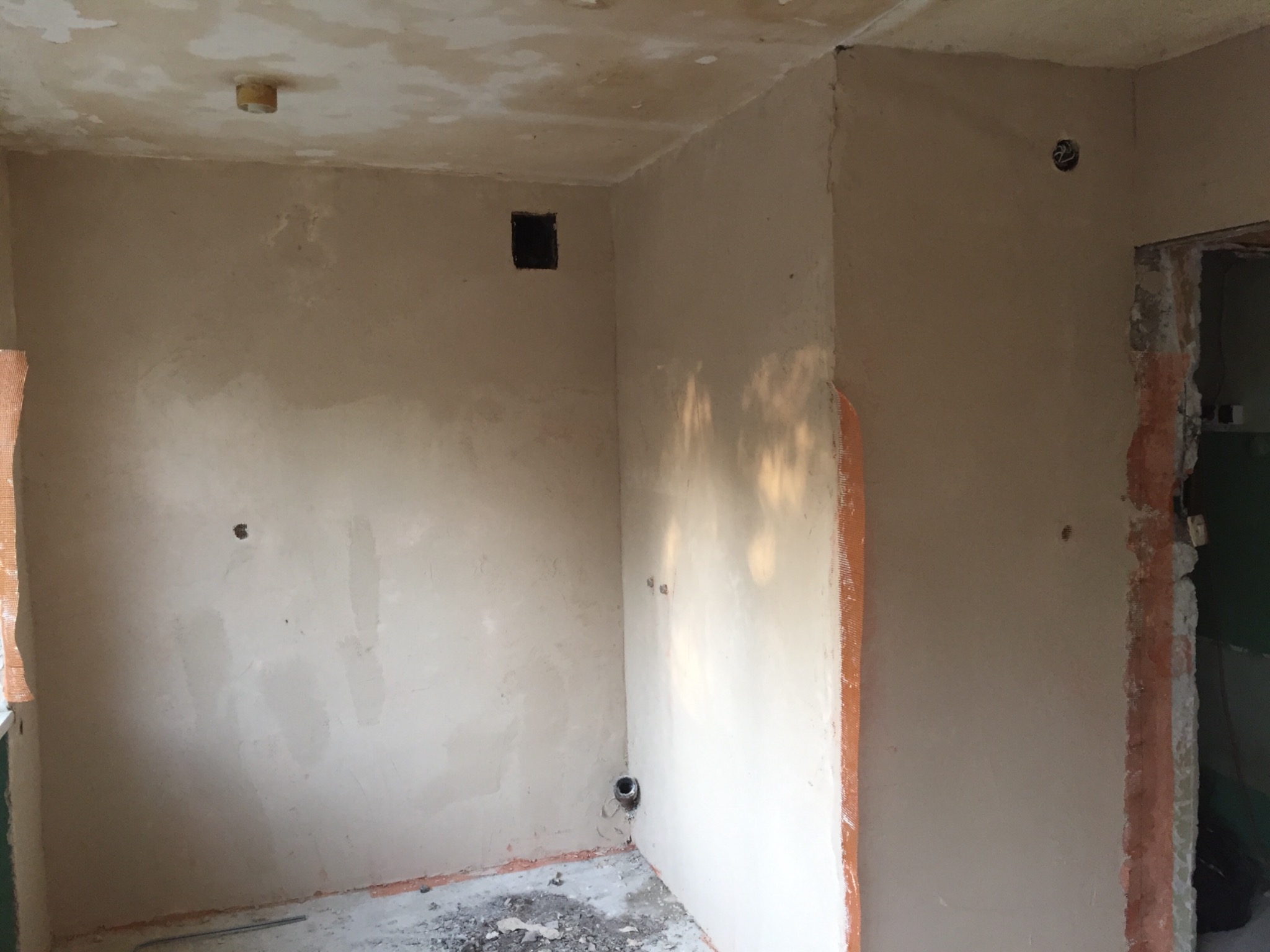 Surowe ściany w pokoju podczas prac przygotowawczych, widoczne ślady po instalacjach, brak wykończenia, resztki gruzu na podłodze, fragment okna z siatką wzmacniającą.