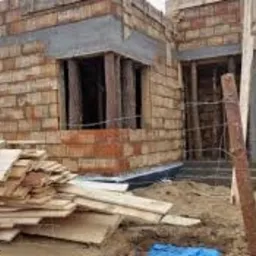 Wznoszenie ścian zewnętrznych domu jednorodzinnego z bloczków ceramicznych, widoczne drewniane stemple podtrzymujące nadproża okienne, na pierwszym planie stos desek i gruz budowlany.