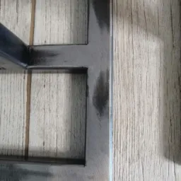 Fragment metalowej konstrukcji z widocznymi śladami piaskowania, leżącej na drewnopodobnej powierzchni.