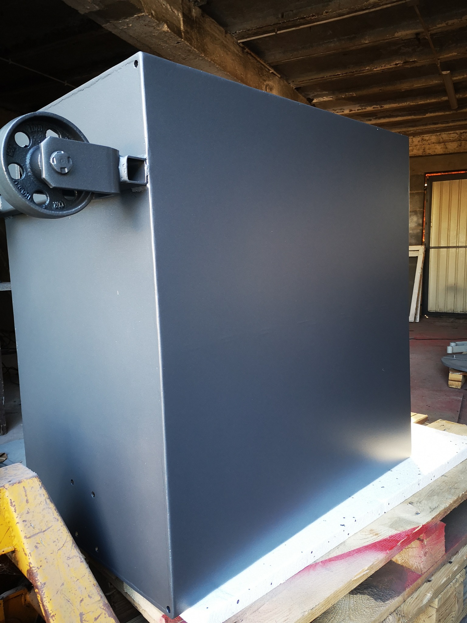 Szary metalowy sześcian z przymocowanym kołem pasowym, po malowaniu proszkowym, leżący na palecie w warsztacie.