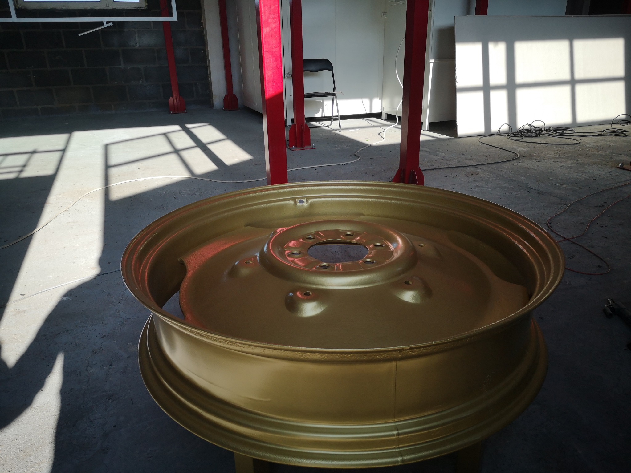 Złota felga samochodowa po renowacji, widoczne ślady po zawieszeniu w procesie malowania, tło warsztatowe z czerwonymi słupami i grą świateł.