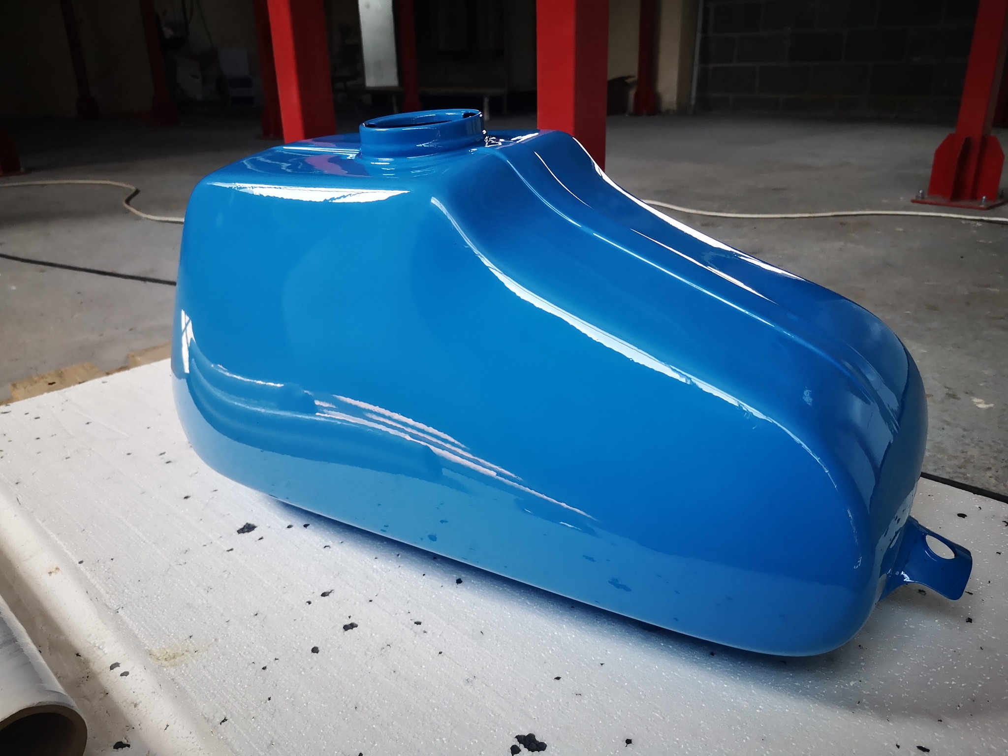 Błękitny, świeżo pomalowany zbiornik paliwa motocykla, leżący na białej powierzchni w warsztacie, z widocznymi elementami konstrukcji budynku w tle.
