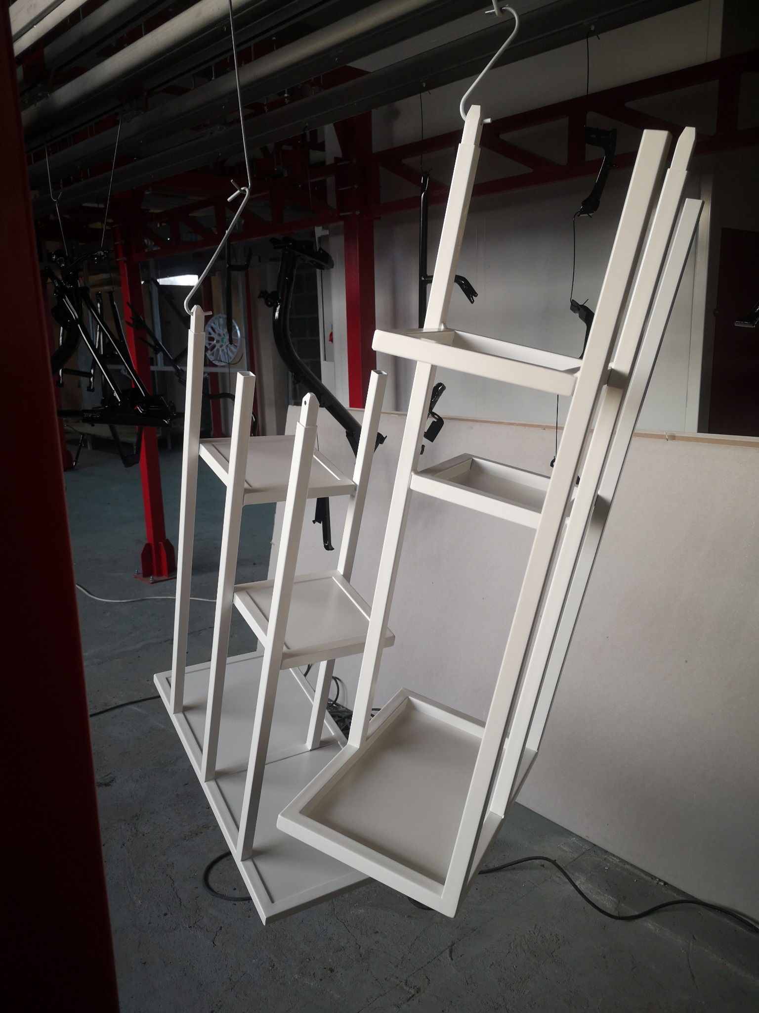 Dwie metalowe konstrukcje regałów z półkami, zawieszone na hakach w hali produkcyjnej, pomalowane na biało, w tle elementy konstrukcji lakierniczej i inne metalowe detale w czarnym kolorze.