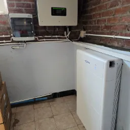 Zestaw fotowoltaiczny SOFAR: inwerter, rozdzielnica i bateria na tle ceglanej ściany. Instalacja w pomieszczeniu z białymi rurami i kafelkami na podłodze.