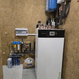 Nowoczesny, biały piec gazowy Calla Verde z podłączeniami hydraulicznymi i elektrycznymi na tle ściany o fakturze baranka. Instalacja w pomieszczeniu technicznym.