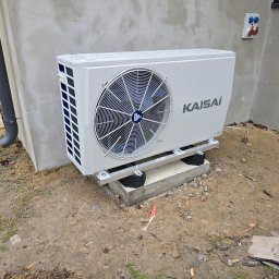 Rami - Patryk Pasieczny - Biała pompa ciepła Kaisai zamontowana na betonowym fundamencie przy ścianie budynku z widocznymi gniazdami elektrycznymi. Widoczny fragment rynny.