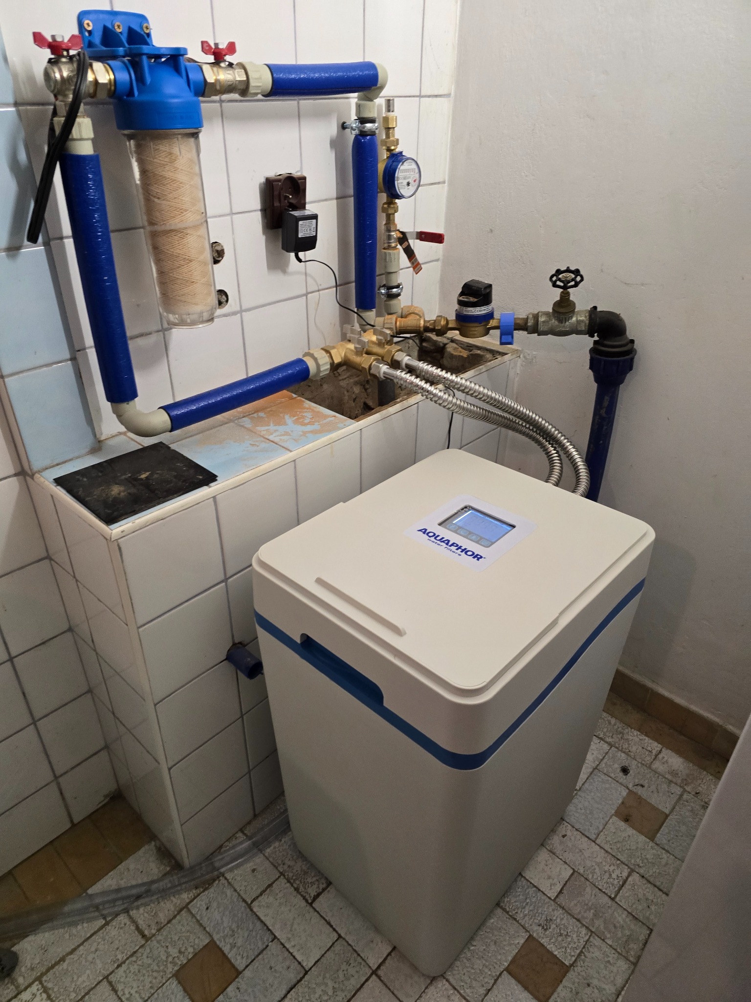 Filtr do wody Aquaphor z niebieskimi rurami w łazience w Zamościu. Widoczny manometr i zawory. Filtr ustawiony na podłodze z płytek.