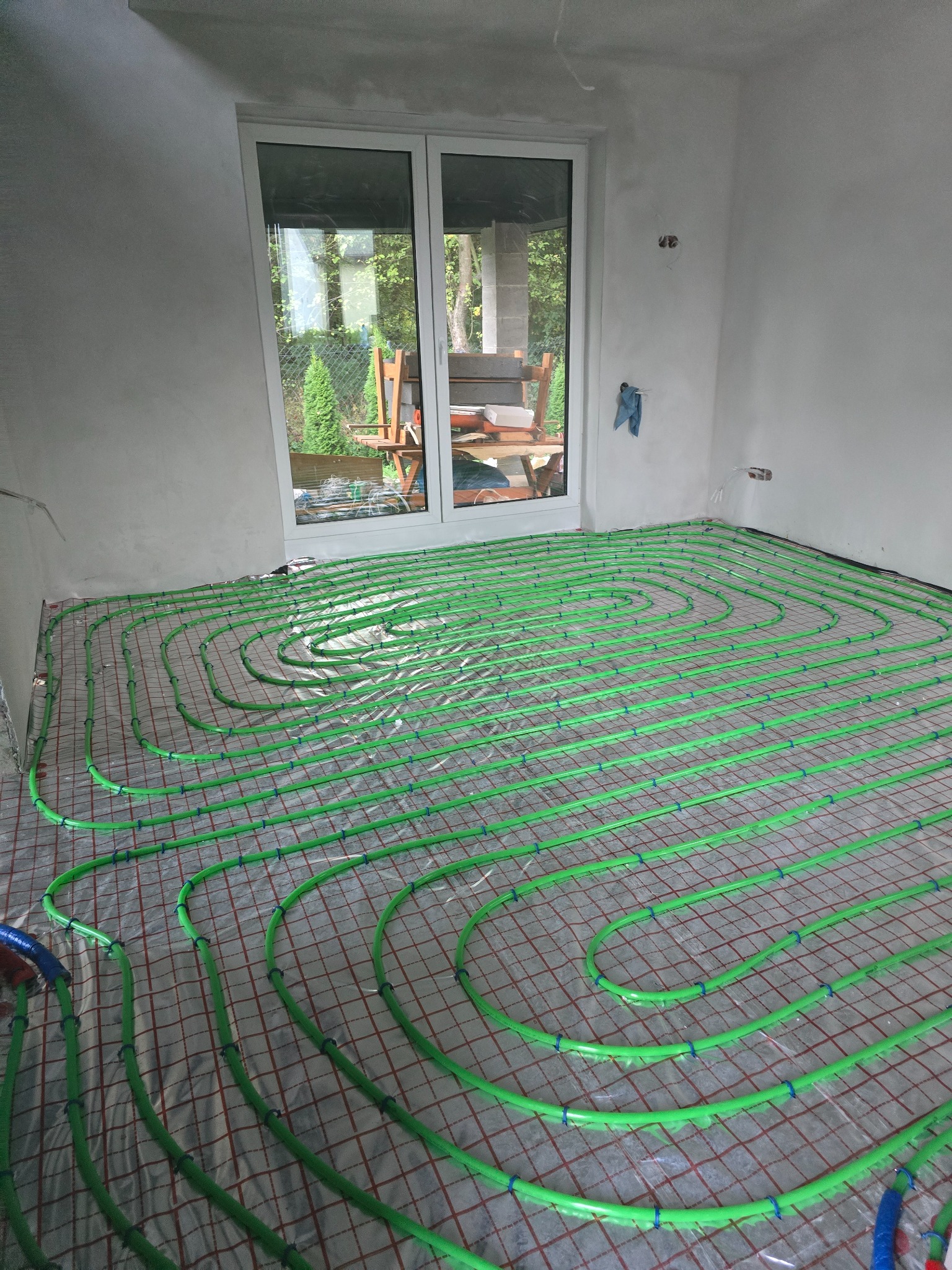 Zielone rury ogrzewania podłogowego ułożone na folii w surowym pomieszczeniu z oknem balkonowym, widok z góry.