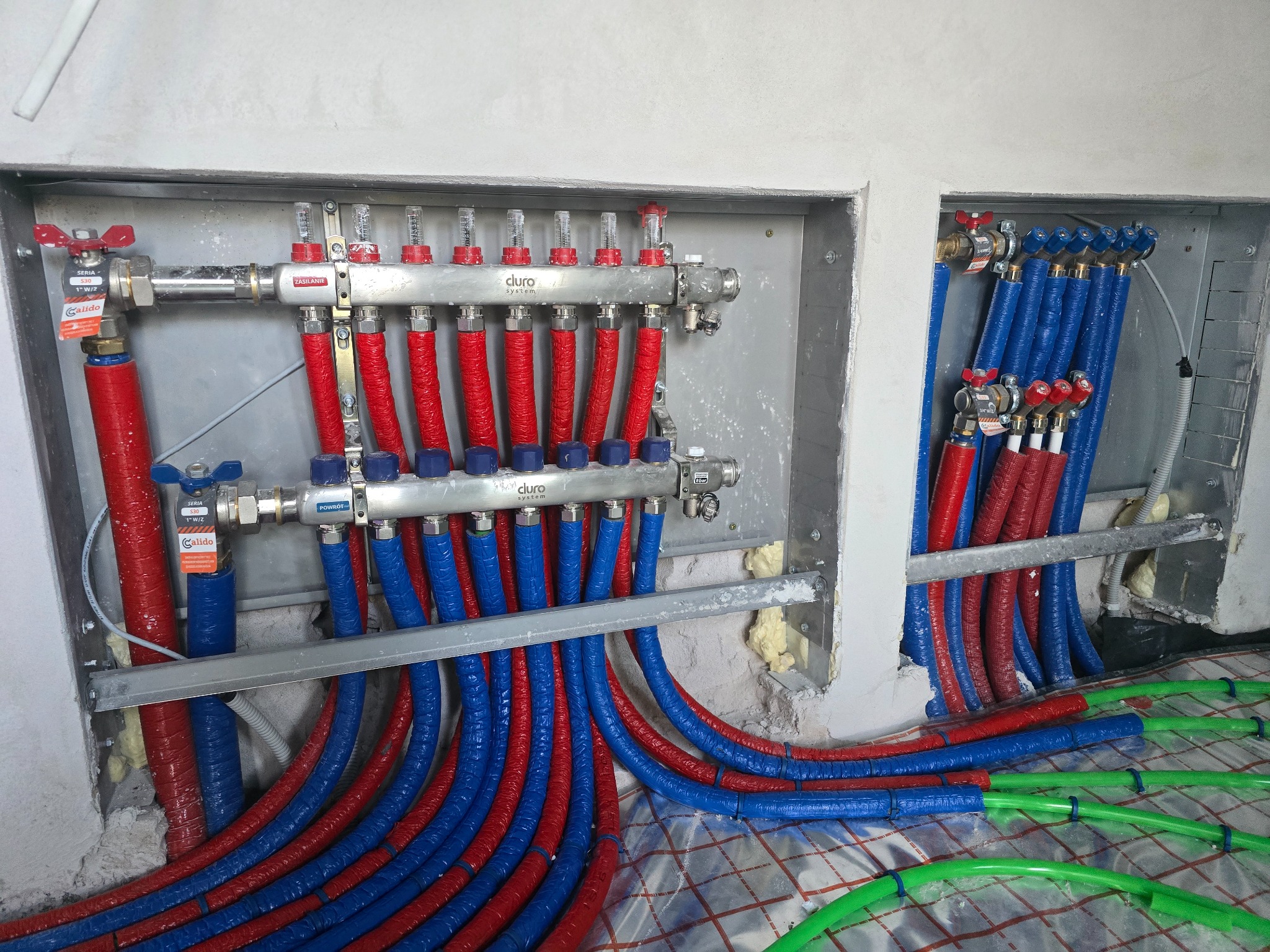 Rozdzielacz ogrzewania podłogowego z czerwono-niebieskimi rurami w izolacji, zamontowany w ścianie, z widoczną zieloną instalacją na podłodze.
