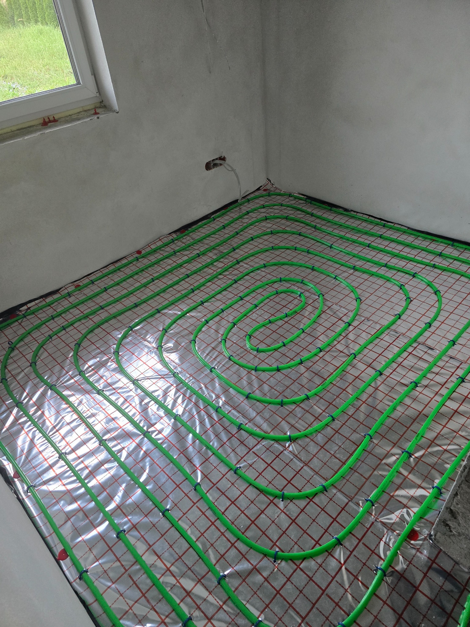 Zielone rury ogrzewania podłogowego ułożone spiralnie na folii z czerwoną siatką w surowym wnętrzu. Widoczny fragment okna i ściany przed wykończeniem.