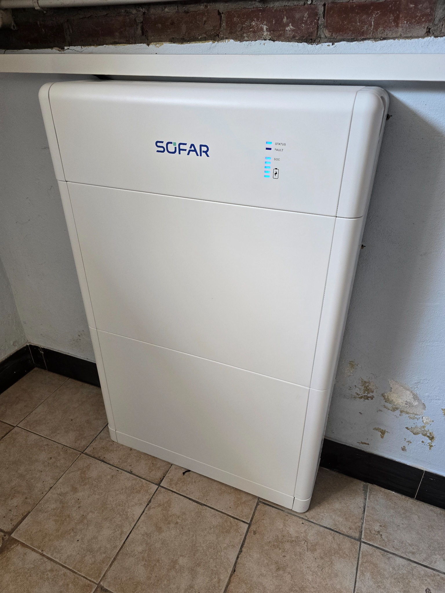 Biały akumulator energii Sofar z niebieskimi diodami LED w pomieszczeniu z kafelkami podłogowymi i ścianą z śladami farby. Widoczny fragment ceglanego muru.