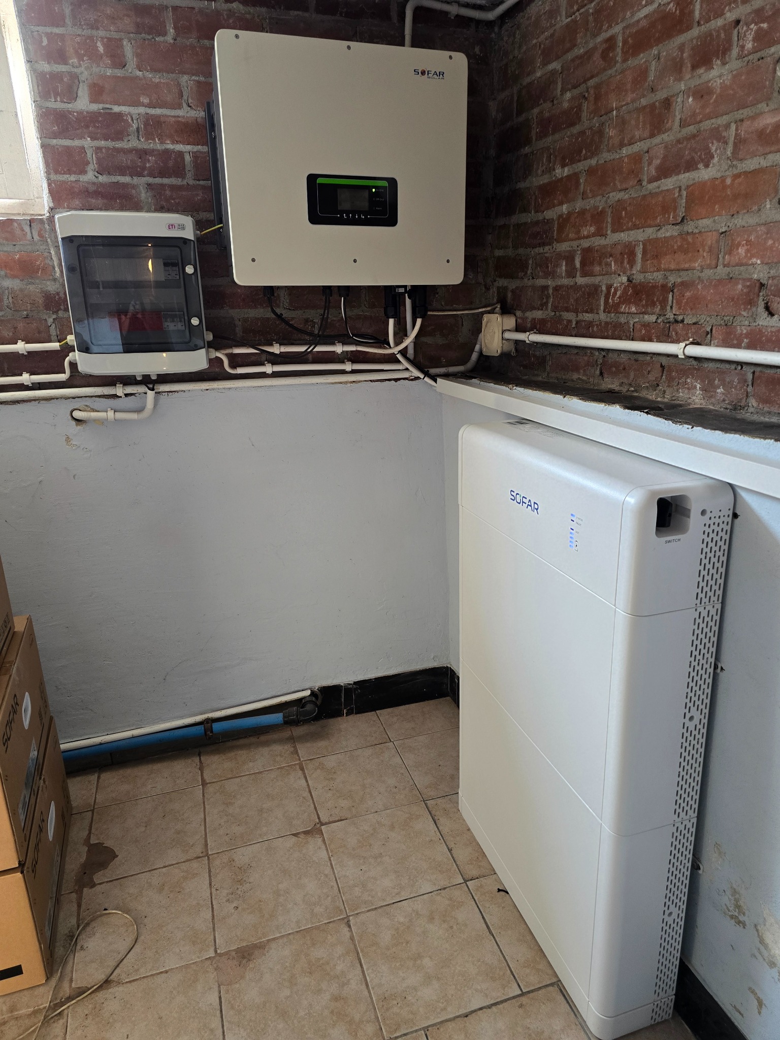 Zestaw fotowoltaiczny SOFAR: inwerter, rozdzielnica i bateria na tle ceglanej ściany. Instalacja w pomieszczeniu z białymi rurami i kafelkami na podłodze.