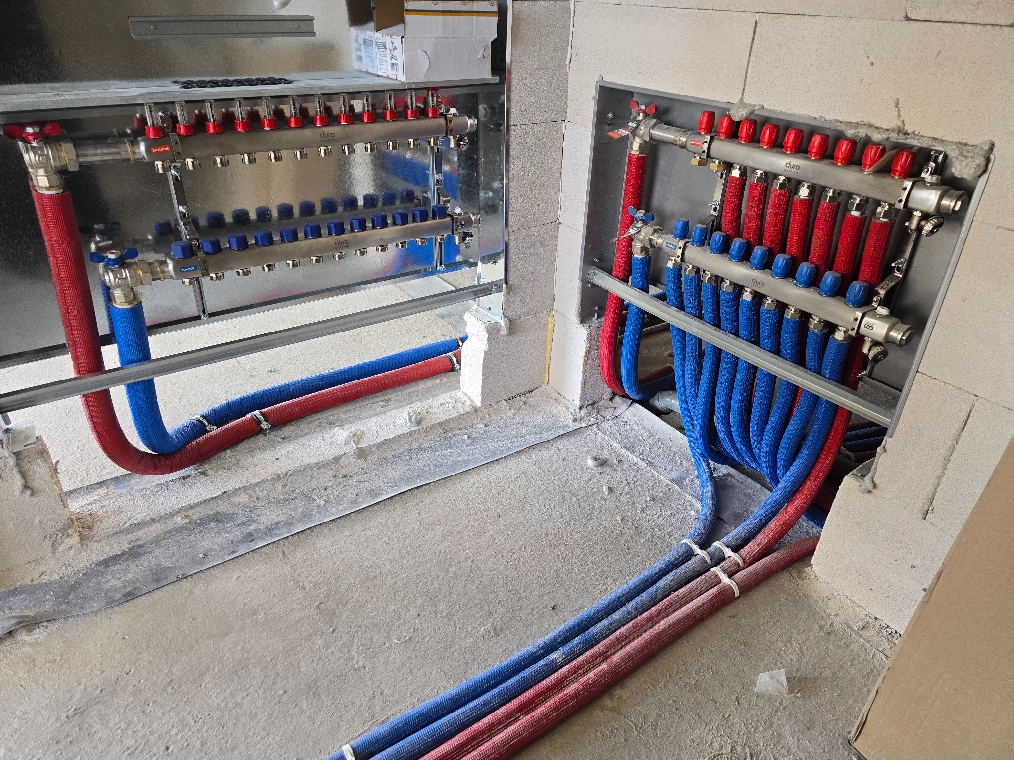 Rozdzielacze ogrzewania podłogowego Duro System zamontowane w ścianie z bloczków, z widocznymi rurami w kolorze niebieskim i czerwonym, ułożonymi w pętle na posadzce.