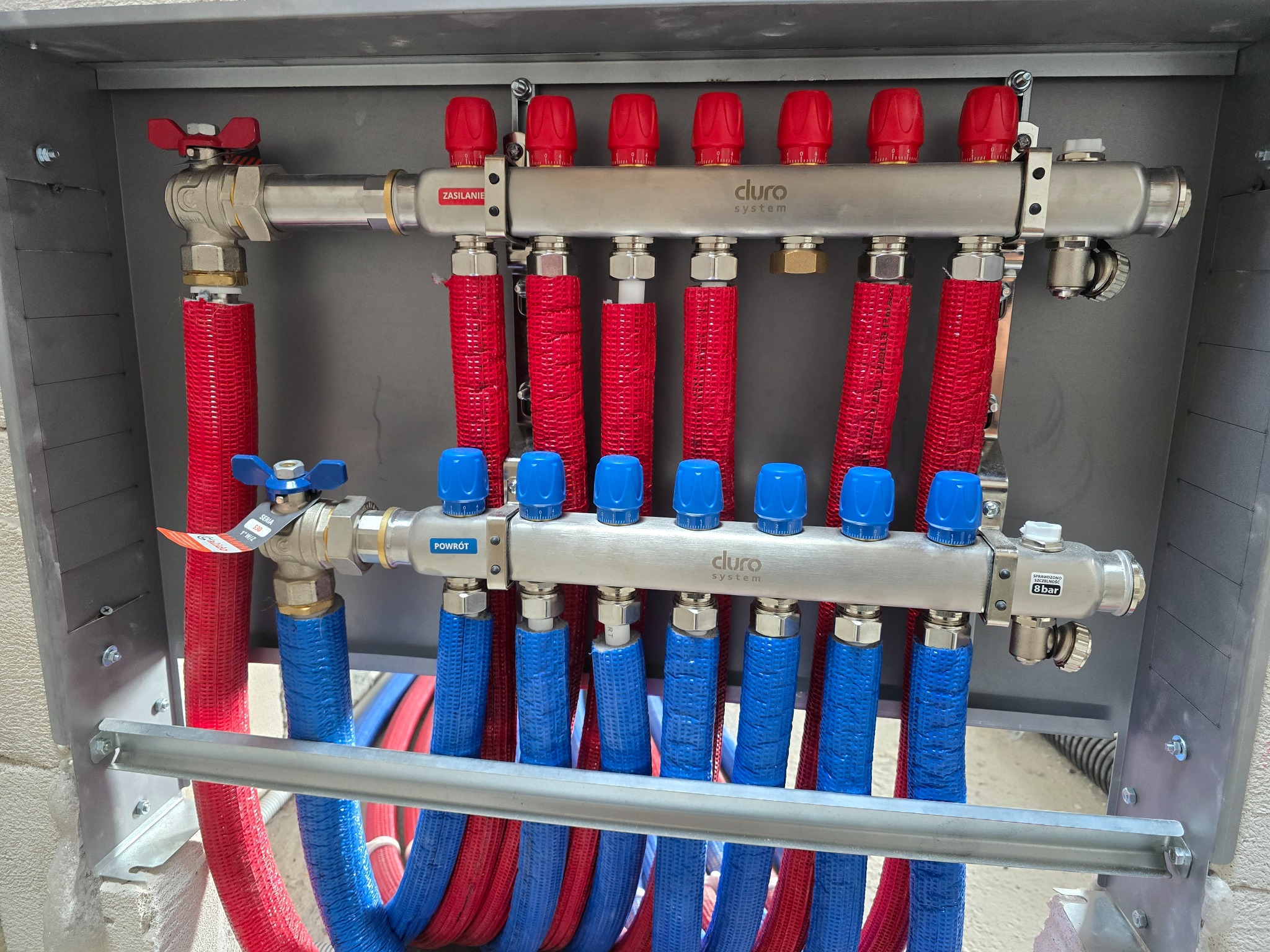 Rozdzielacz ogrzewania podłogowego Cluro System w szarej szafce, z czerwonymi rurami zasilającymi i niebieskimi powrotnymi. Widoczne zawory i oznaczenia 'Zasilanie' i 'Powrót'.